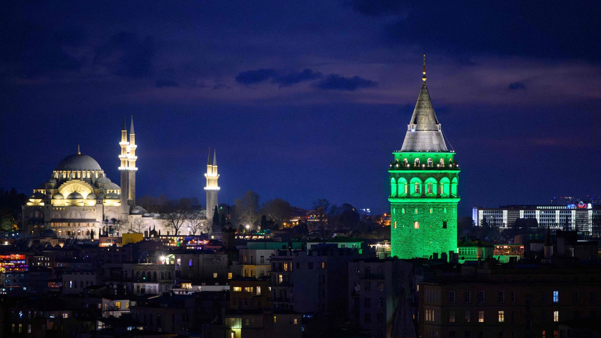 Grün angestrahlter Galata-Turm mit Moschee im Hintergrund bei Nacht.