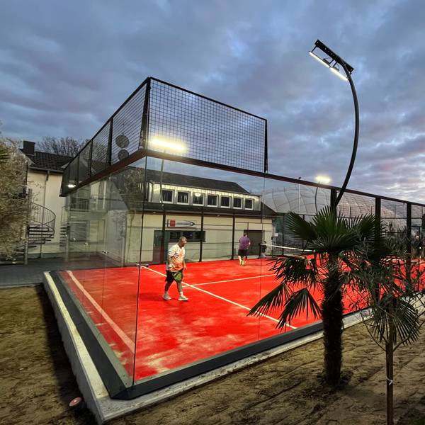 Eine Art Mini-Tennisplatz mit Glaswänden steht vor einem Vereinsheim.