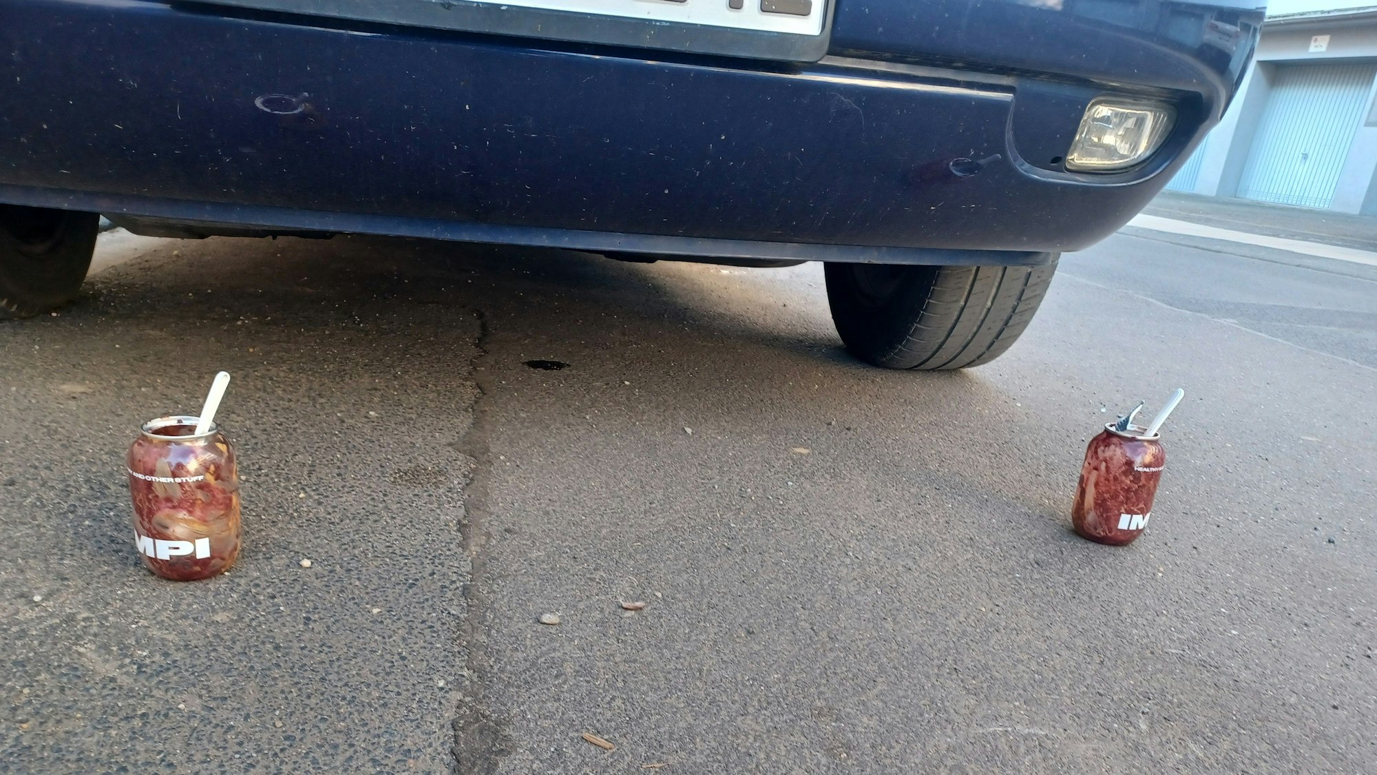 Zwei leere Dosen mit Impi-Aufdruck stehen hinter einem geparkten Auto.