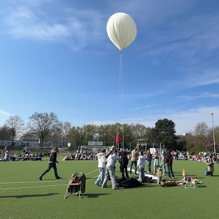 Jugendliche stehen auf einem Sportplatz um einen weißen Ballon herum.