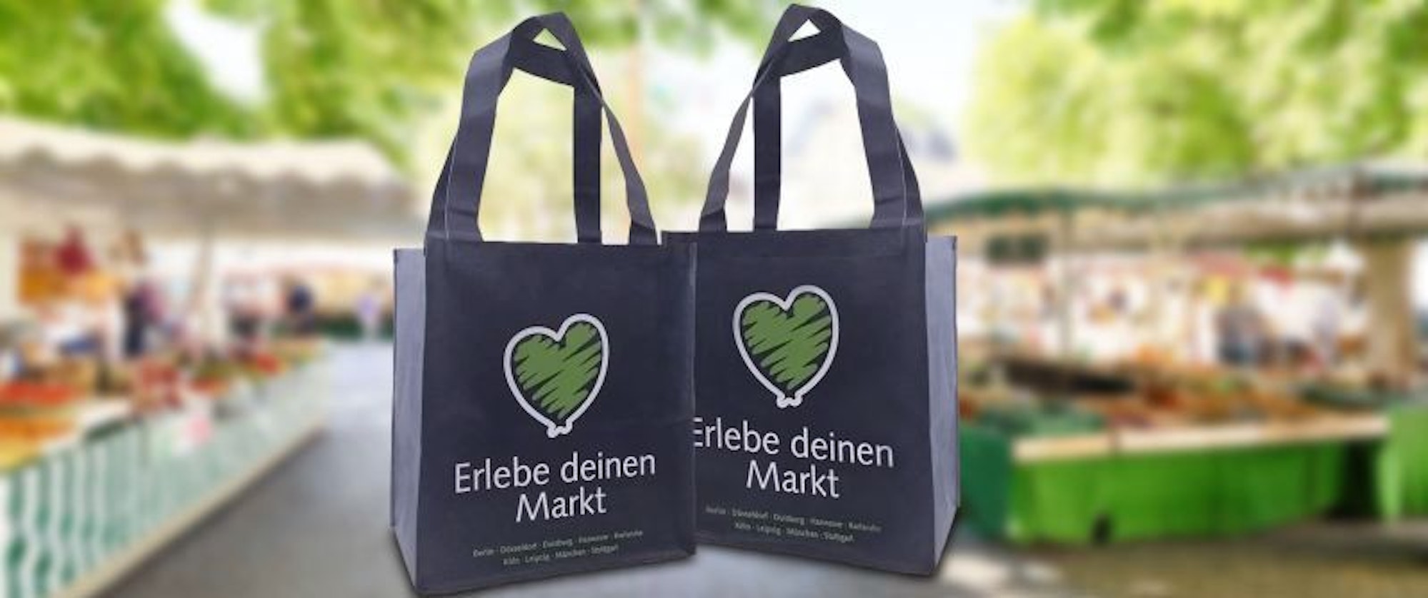 Zwei graue Einkaufstaschen mit dem Schriftzug "Erlebe deinen Markt" vor einem unscharfen Hintergrund, der einen Wochenmarkt zeigt.