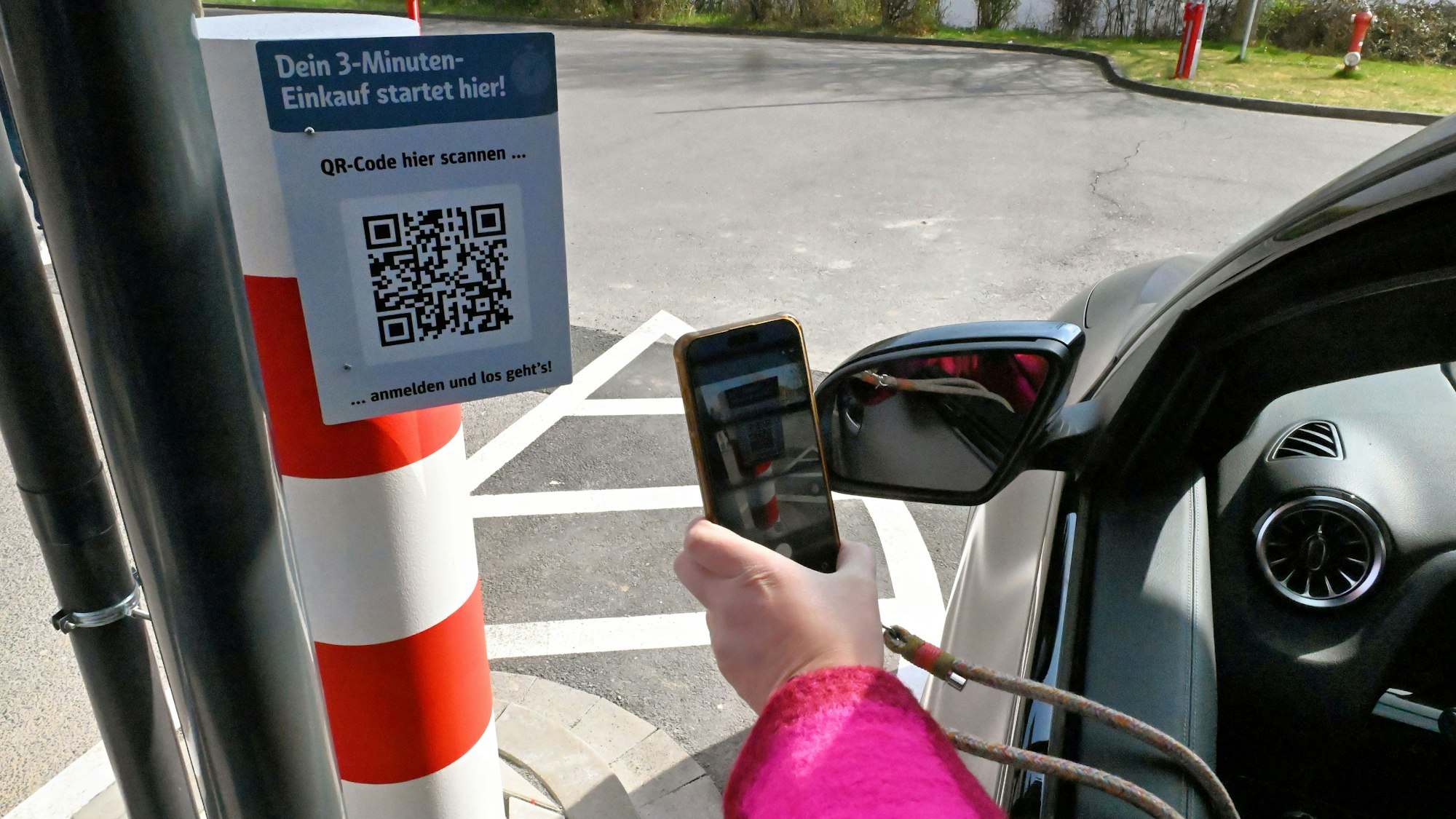 Aus einem Autofenster ist die Hand einer Frau zu sein, die ein Smartphone hält und einen QR-Code scannt.