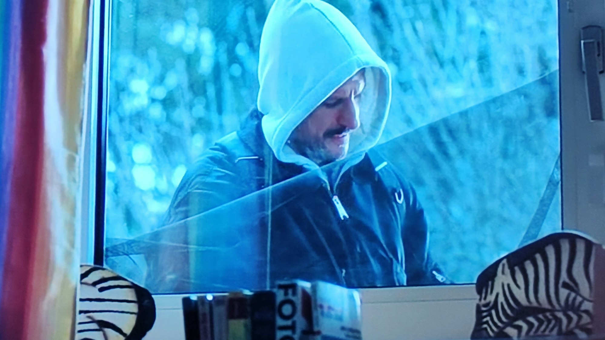 Ein Mann mit einer Kapuze auf dem Kpf ist dabei, ein Fenster aufzubrechen. Die Filmszene zeigt die Perspektive aus dem Wohnraum. Im Vordergrund stehen Dekostückle auf der Fensterbank.