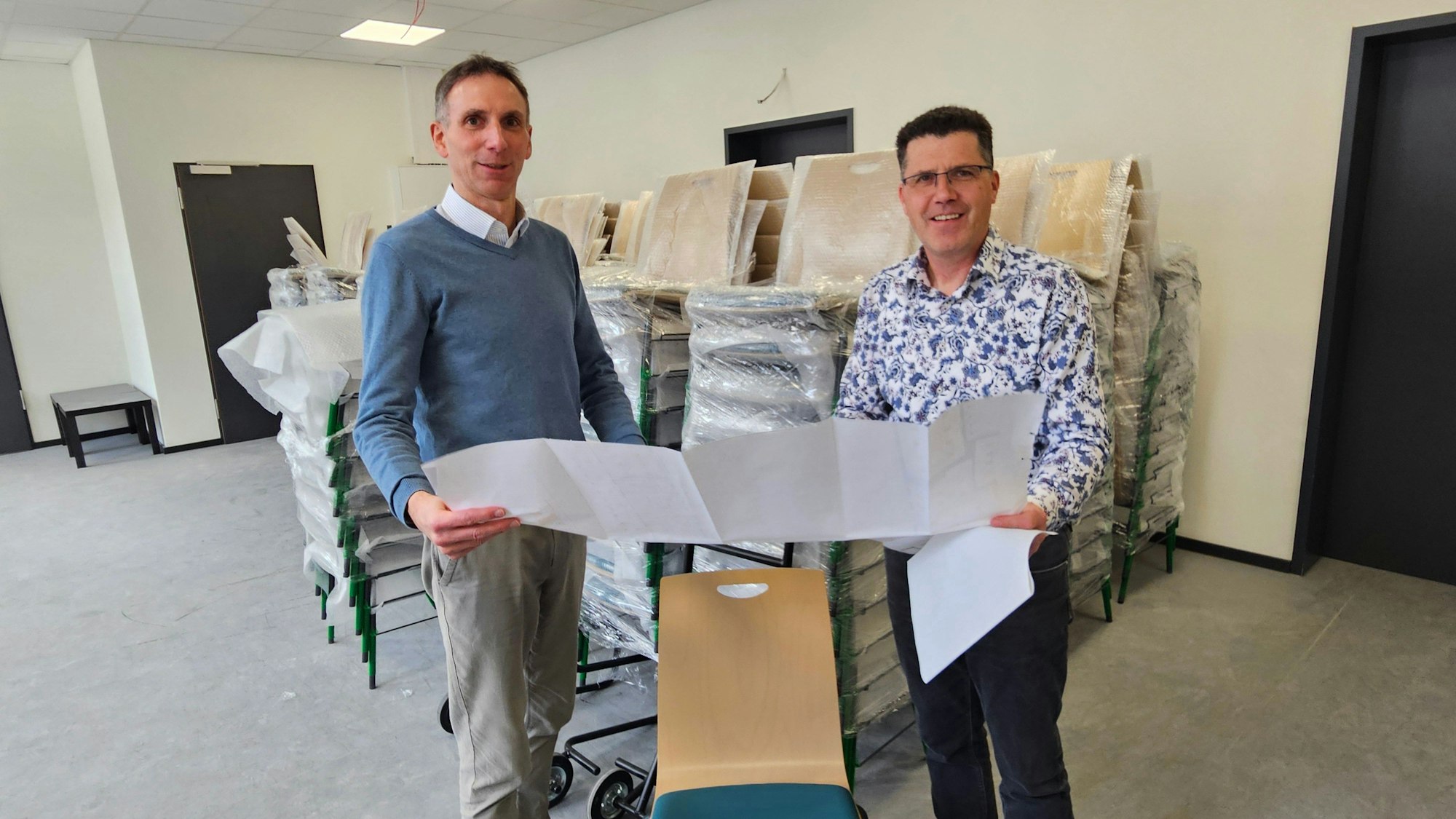 Zwei Männer stehen nebeneinander und halten eine Planskizze fest. Im Hintergrund sind Stühle zu sehen.