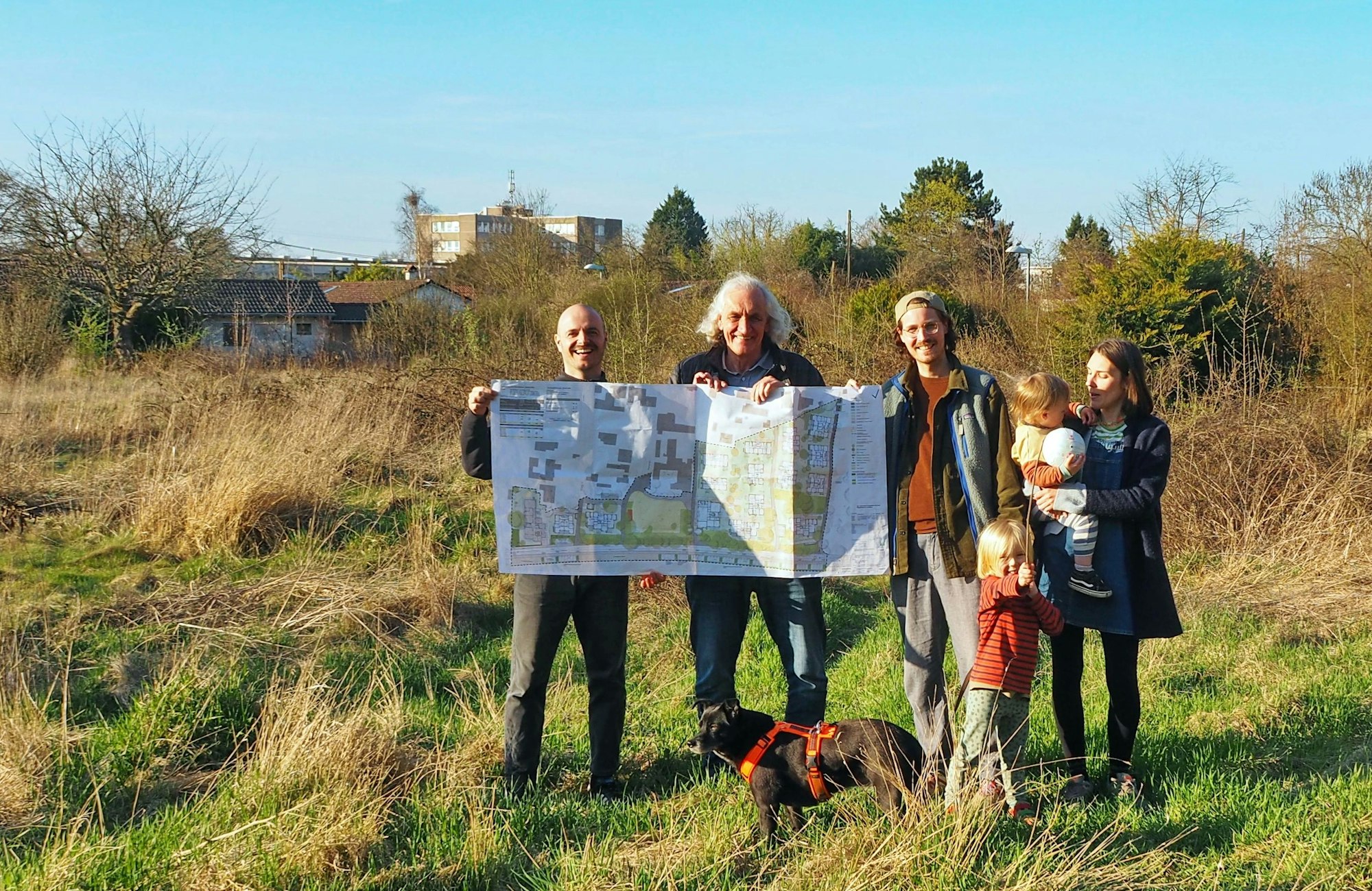 Mehrere Erwachsene, zwei Kinder und ein Hund stehen auf einer Wiese. Zwei Männer halten einen Bauplan.