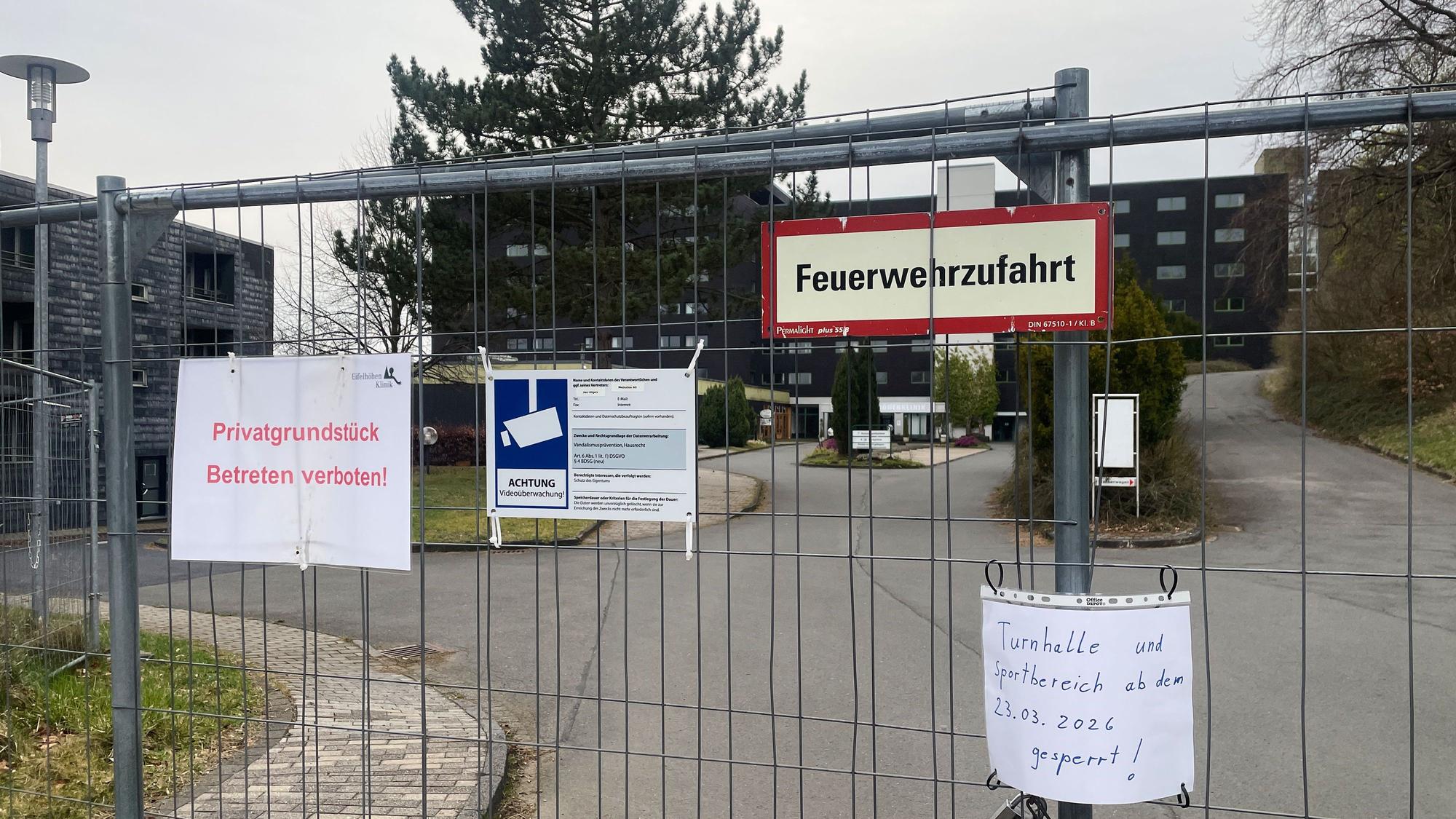 Das Gelände der ehemaligen Eifelhöhen-Klinik ist mit einem Zaun abgesperrt, an dem mehrere Schilder befestigt sind.