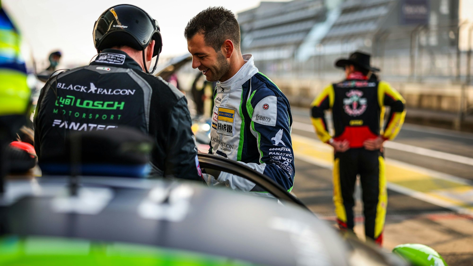 Tobias Müller unterhält sich mit einem anderen Teammitglied in der Boxengasse. Im Hintergrund steht jemand in Rennoverall und Cowboyhut.