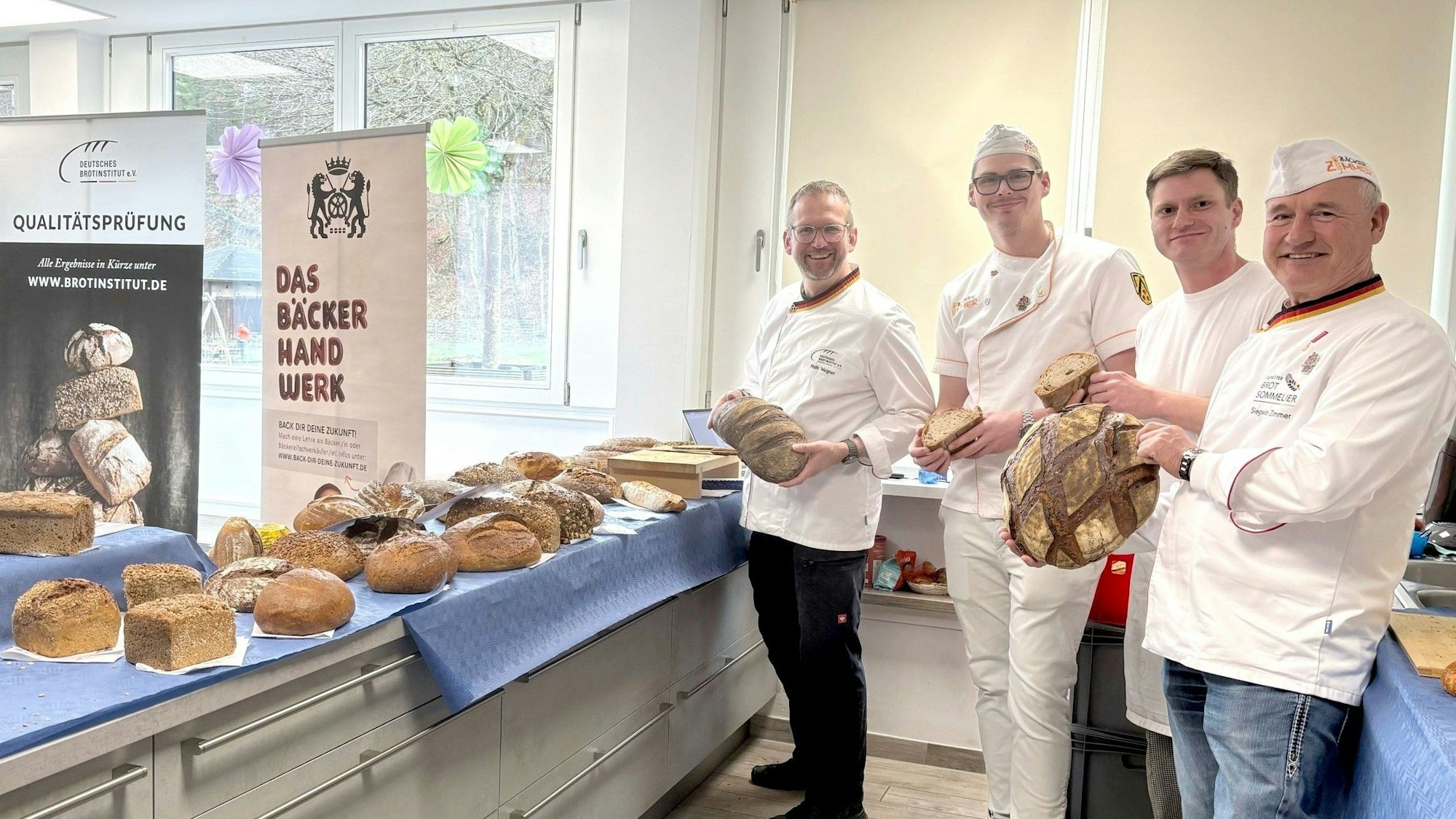 Vier Männer in Bäckerkleidung stehen nebeneinander und halten Brote in ihren Händen.