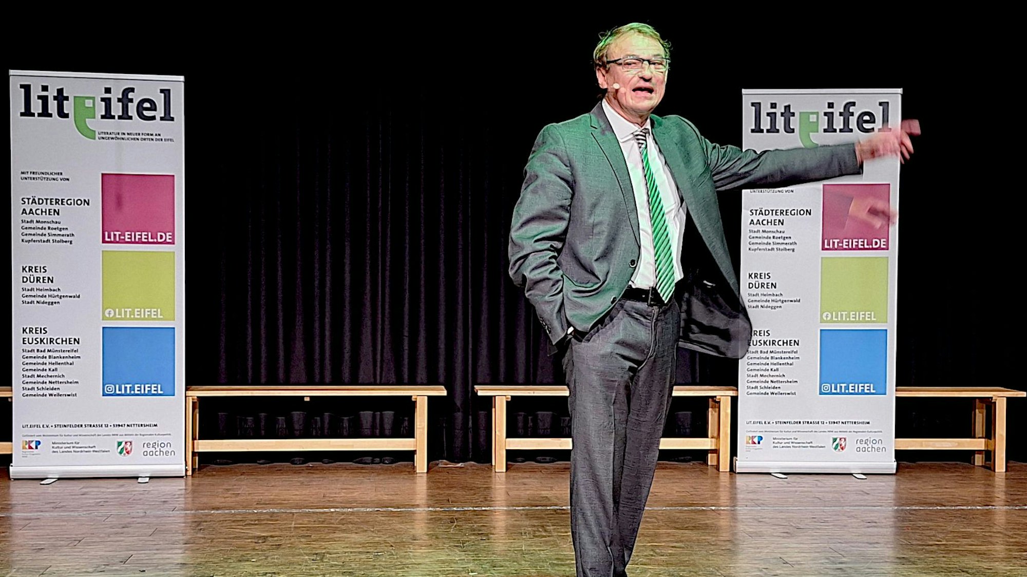 Autor und Papst-Experte Andreas Englisch steht auf einer Bühne und spricht in ein Mikrofon. Im Hintergrund sind zwei Lit.Eifel-Aufsteller zu sehen.