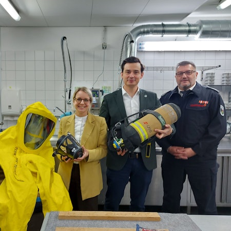 Drei Männer, davon zwei in Feuerwehruniform, und eine Frau im gelben Blazer stehen in einer Werkstatt neben einem gelben Ganzkörperschutzanzug und halten eine Atemschutzmaske und eine Sauerstoffflasche in der Hand.