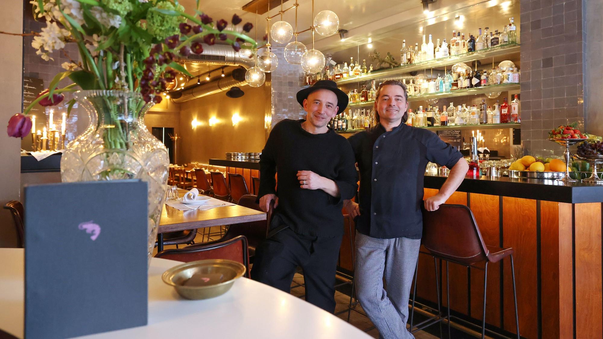 Die beiden Männer stehen vor der Bar und lachen in die Kamera.