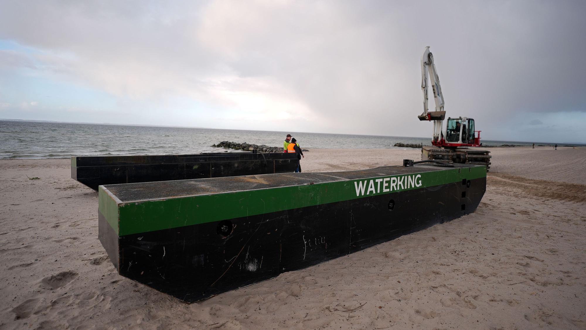 Ein Spezial-Schwimmbagger (r) wird neben Schwimmpontons an den Strand bei Niendorf gefahren, wo ein Wal im flachen Wasser gestrandet ist.