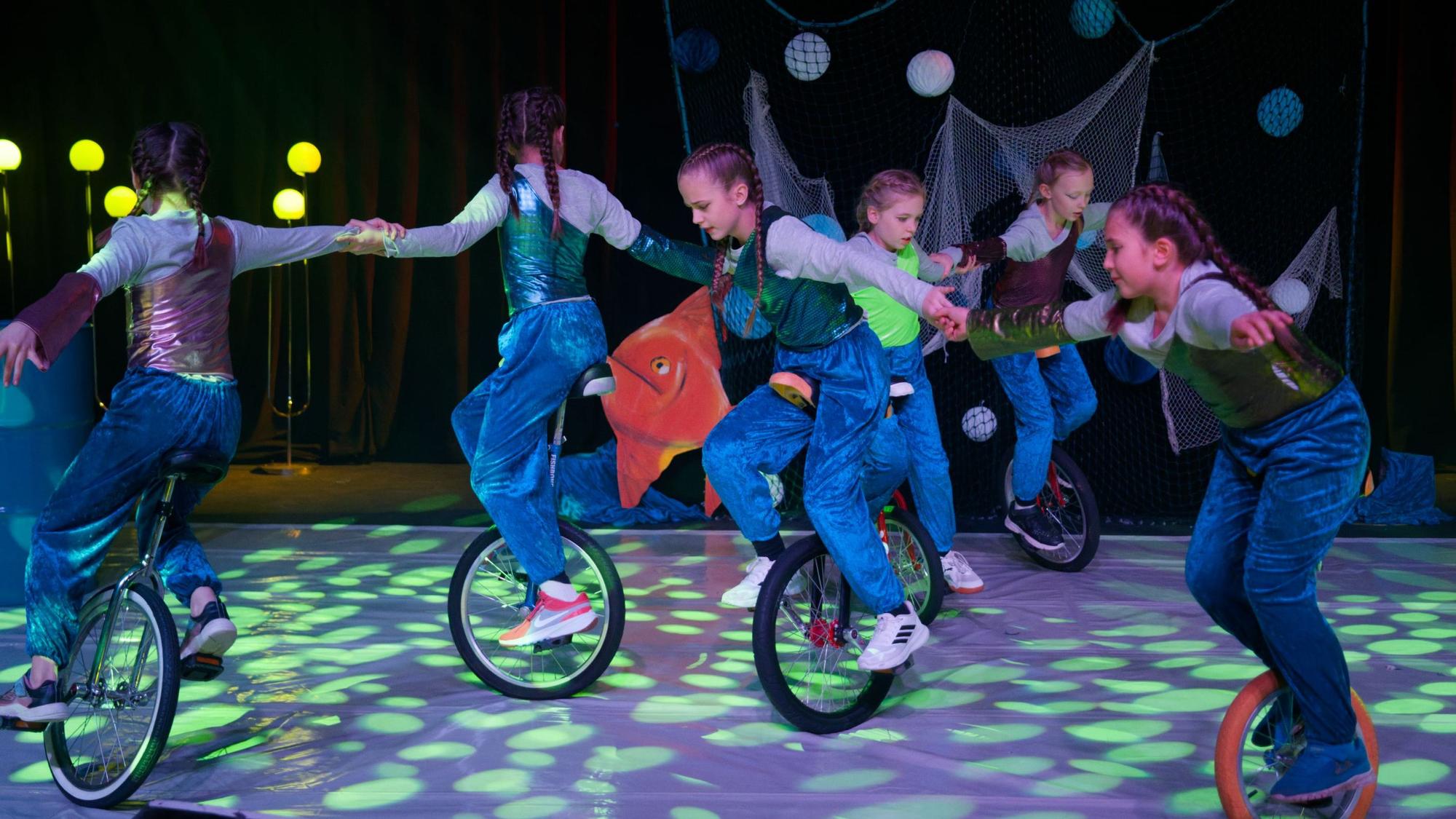 Sechs Mädchen mit geflochtenen Zöpfen fahren in einer bunt beleuchteten Zirkus-Manege auf dem Einrad und halten sich an den Händen.