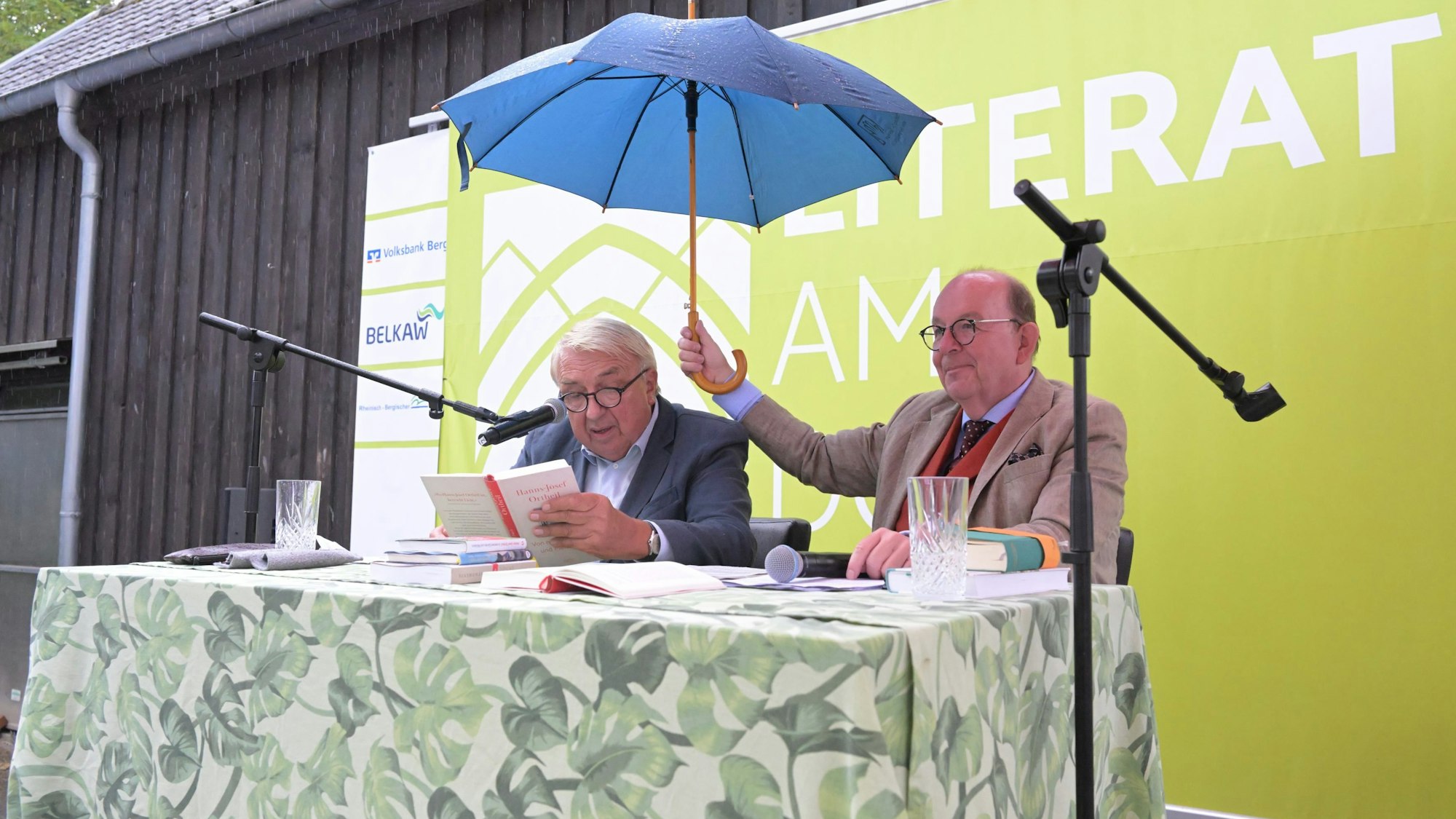 Ein Mann liest auf einer Bühne aus einem Buch. Daneben hält ein Mann einen blauen Regenschirm über ihn.