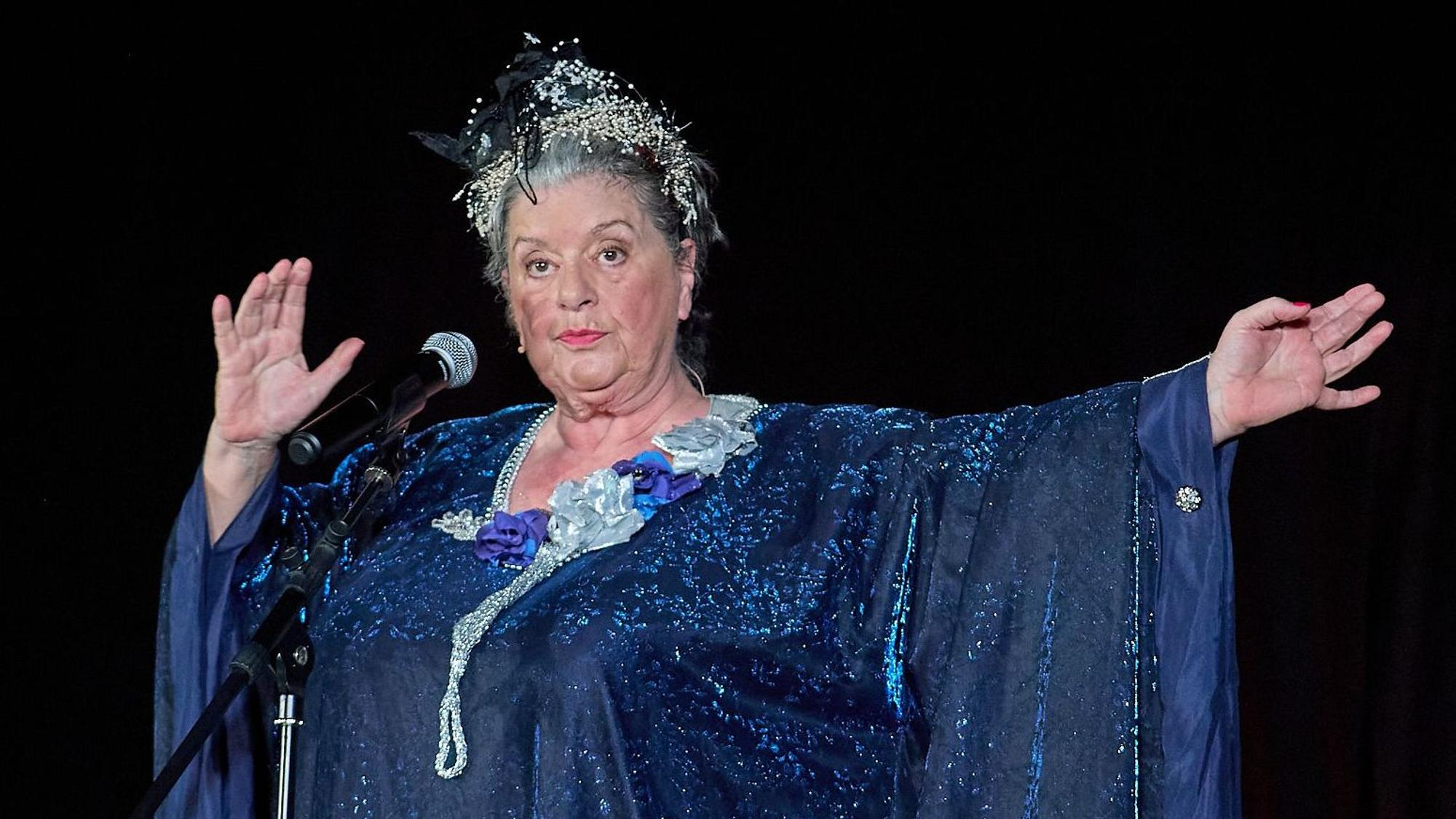 Biggi Wanninger steht in einem blauen Kleid mit Haarschmuck auf der Bühne.