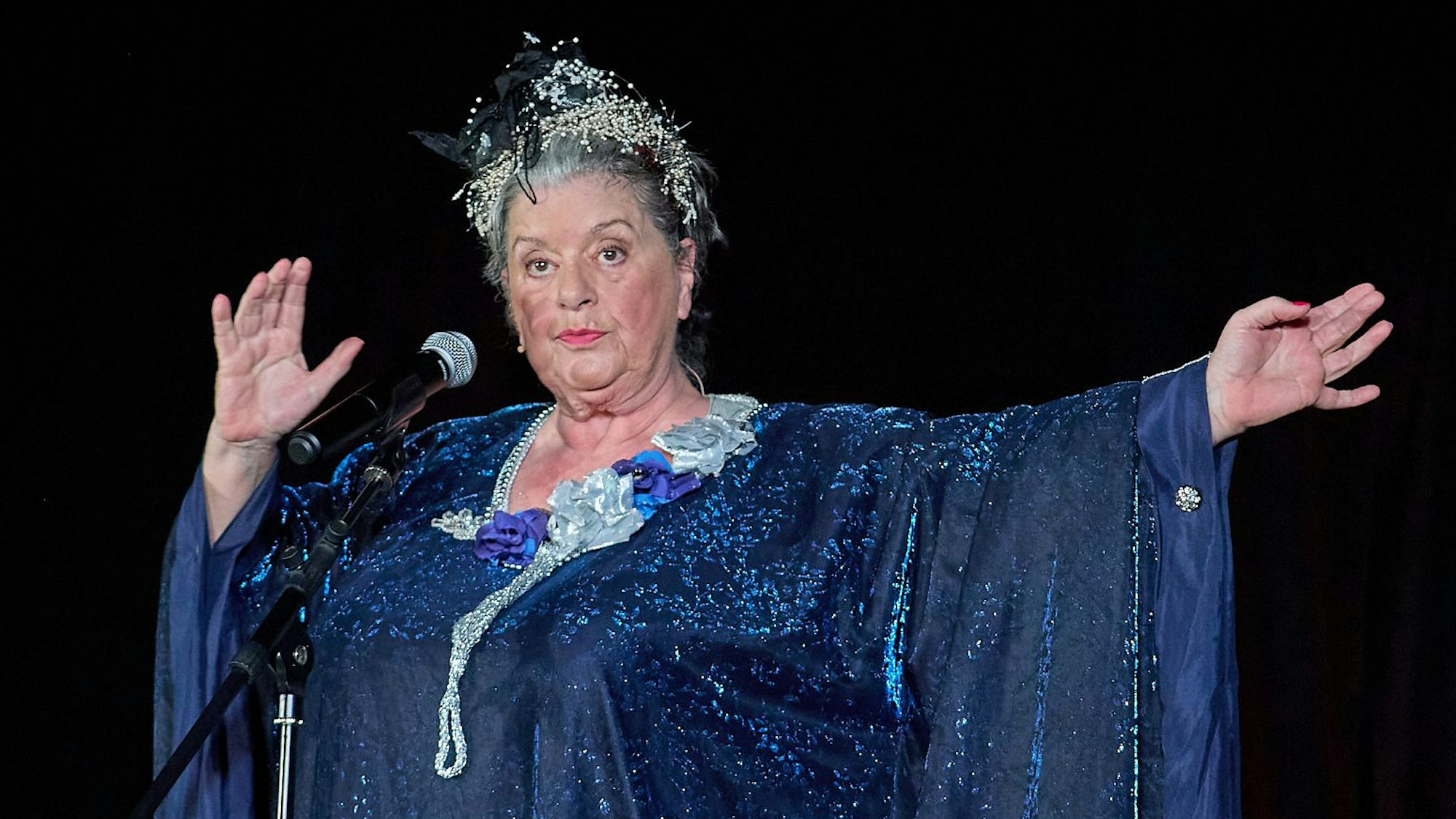 Biggi Wanninger steht in einem blauen Kleid mit Haarschmuck auf der Bühne.