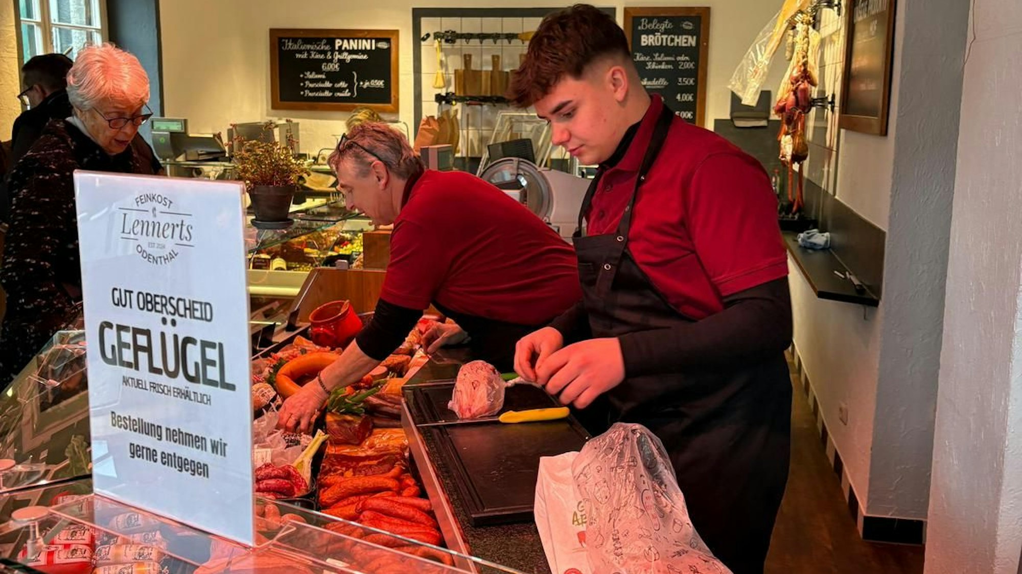 Zwei Metzger stehen hinter einer Fleischtheke und bedienen. Davor mehrere Kunden.