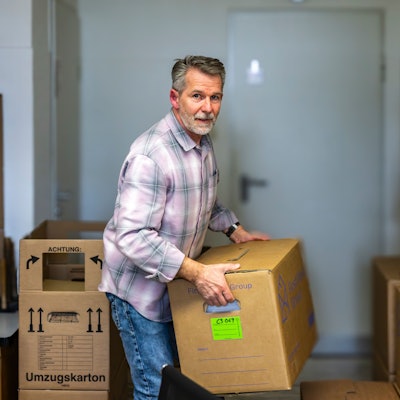 Ein Mann trägt einen Umzugskarton. Weitere Kartons stehen um ihn herum.