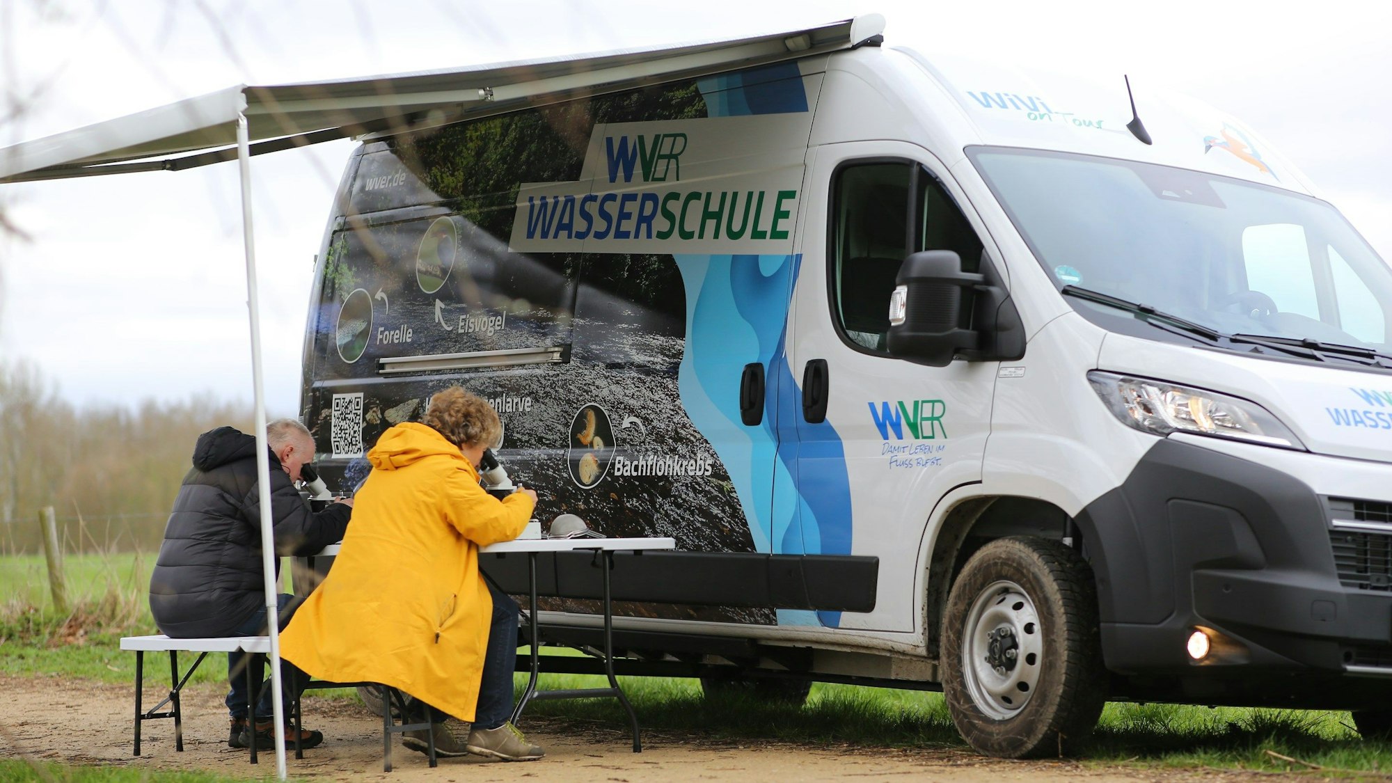 Ein Mann und eine Frau sitzen an einem Tisch unter einer Markise, die an dem Wasserschul-Fahrzeug des WVER befestigt ist. Beide schauen in Mikroskope.