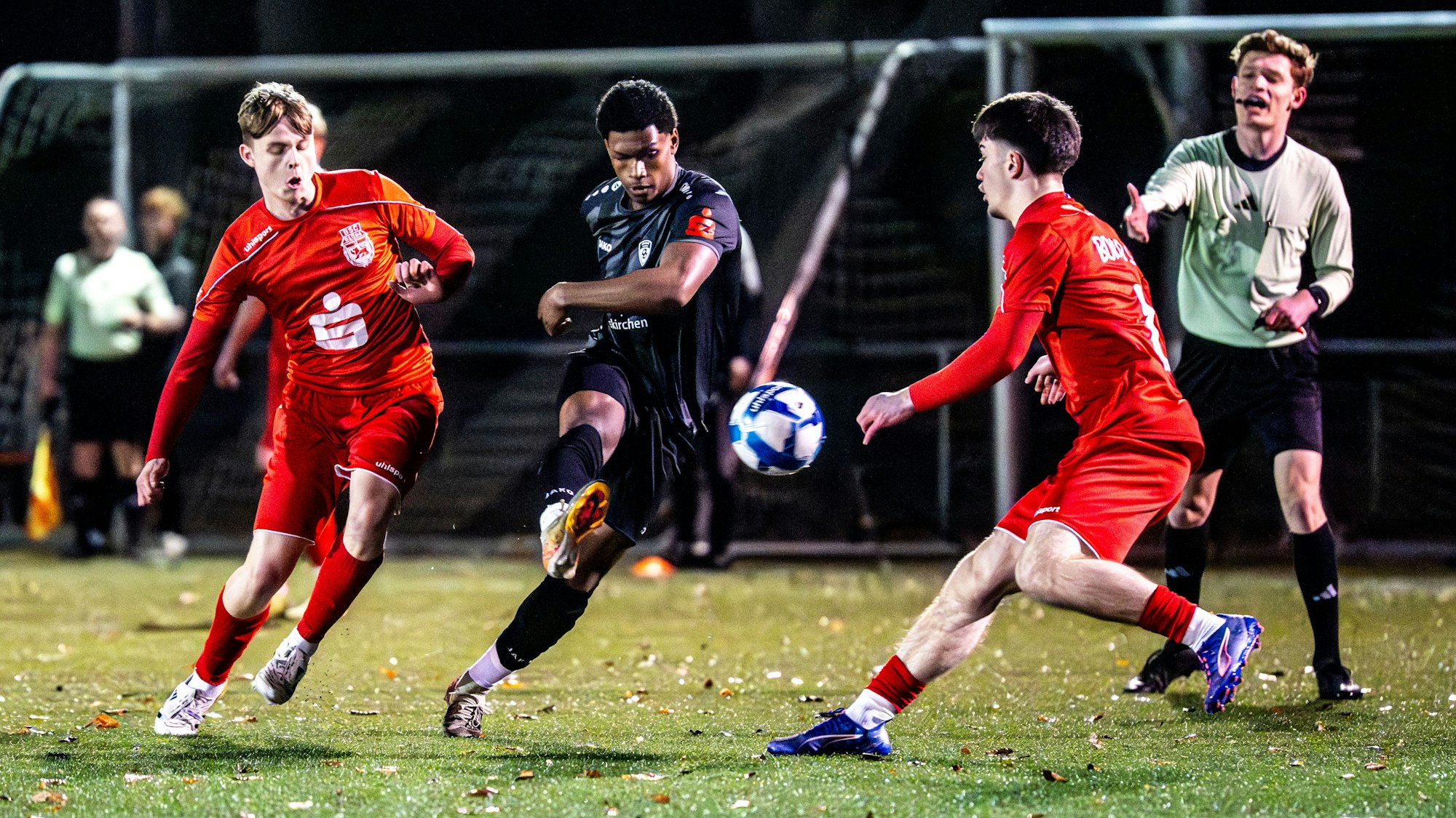 JSG-Spieler Mamadou Diaby (M.) mit dem Ball in die Tiefe. Zwei Gegenspieler bedrängen ihn, im Hintergrund gestikuliert der Schiedsrichter.