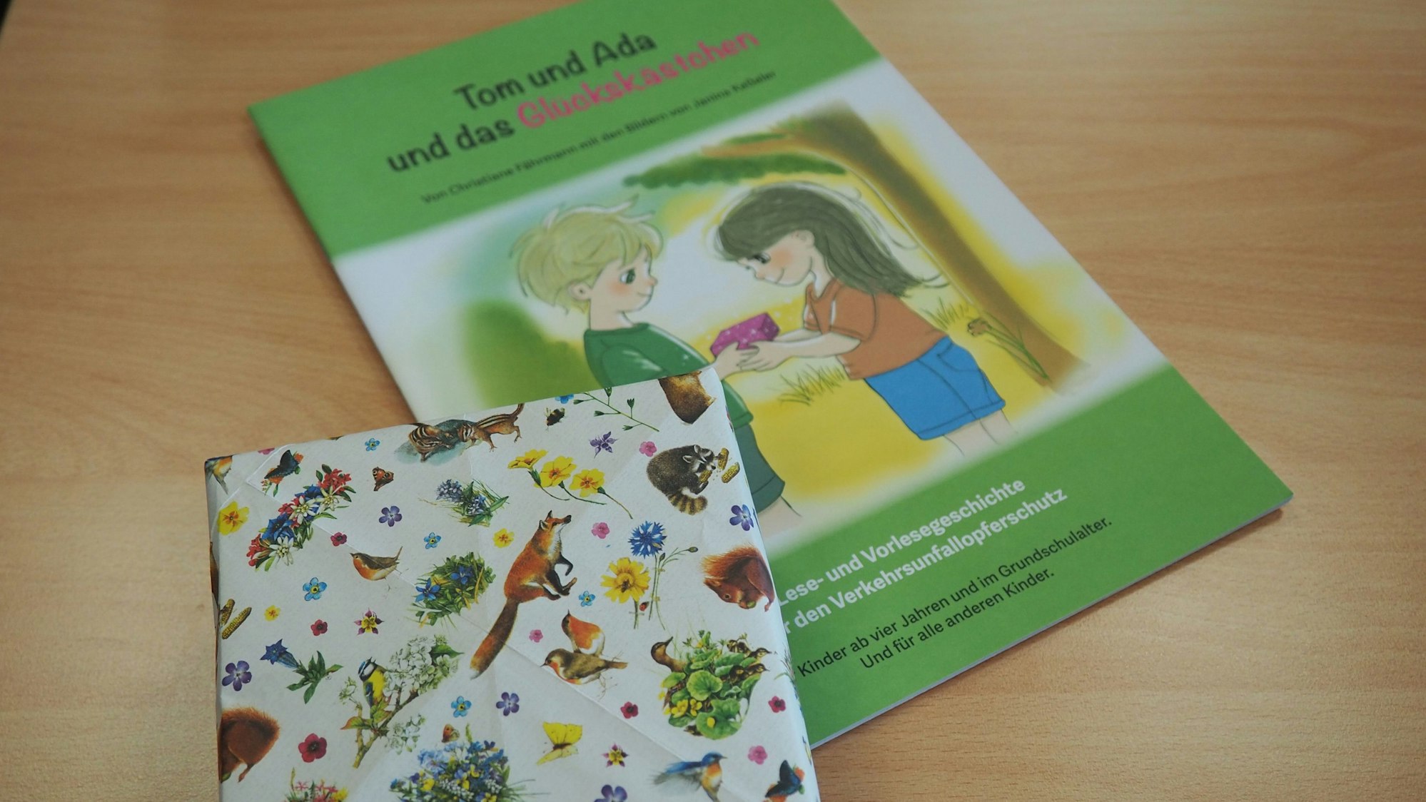 Das Buch „Tom und Ada und das Glückskästchen“ liegt auf einem Tisch, ebenso ein Kästchen aus buntem Geschenkpapier.