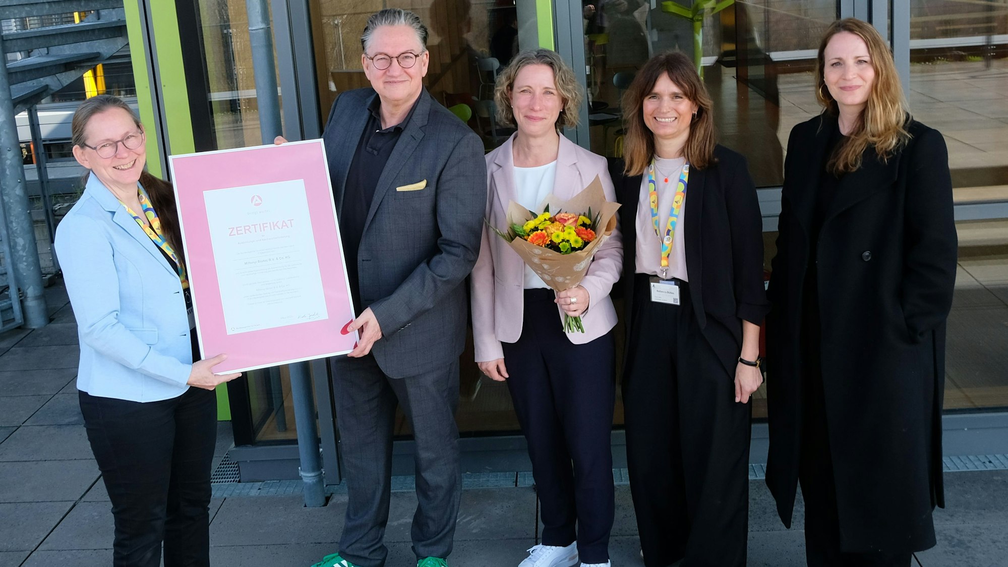 Fünf Personen stehen vor einem Gebäude, halten ein Zertifikat hoch und lächeln.