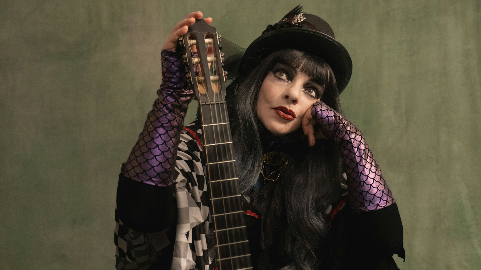 Nina Hagen trägt einen schwarzen Hut mit Feder und stützt sich auf den Hals ihrer Gitarre.