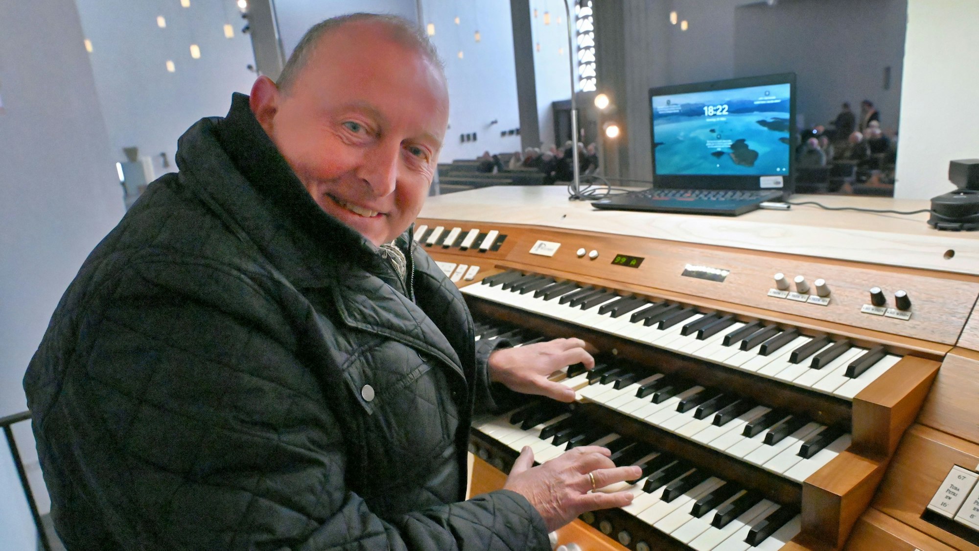 Otto Maria Krämer sitzt am Spieltisch der Orgel, auf dem ein Laptop steht. Der Organist hat eine dicke Jacke an.