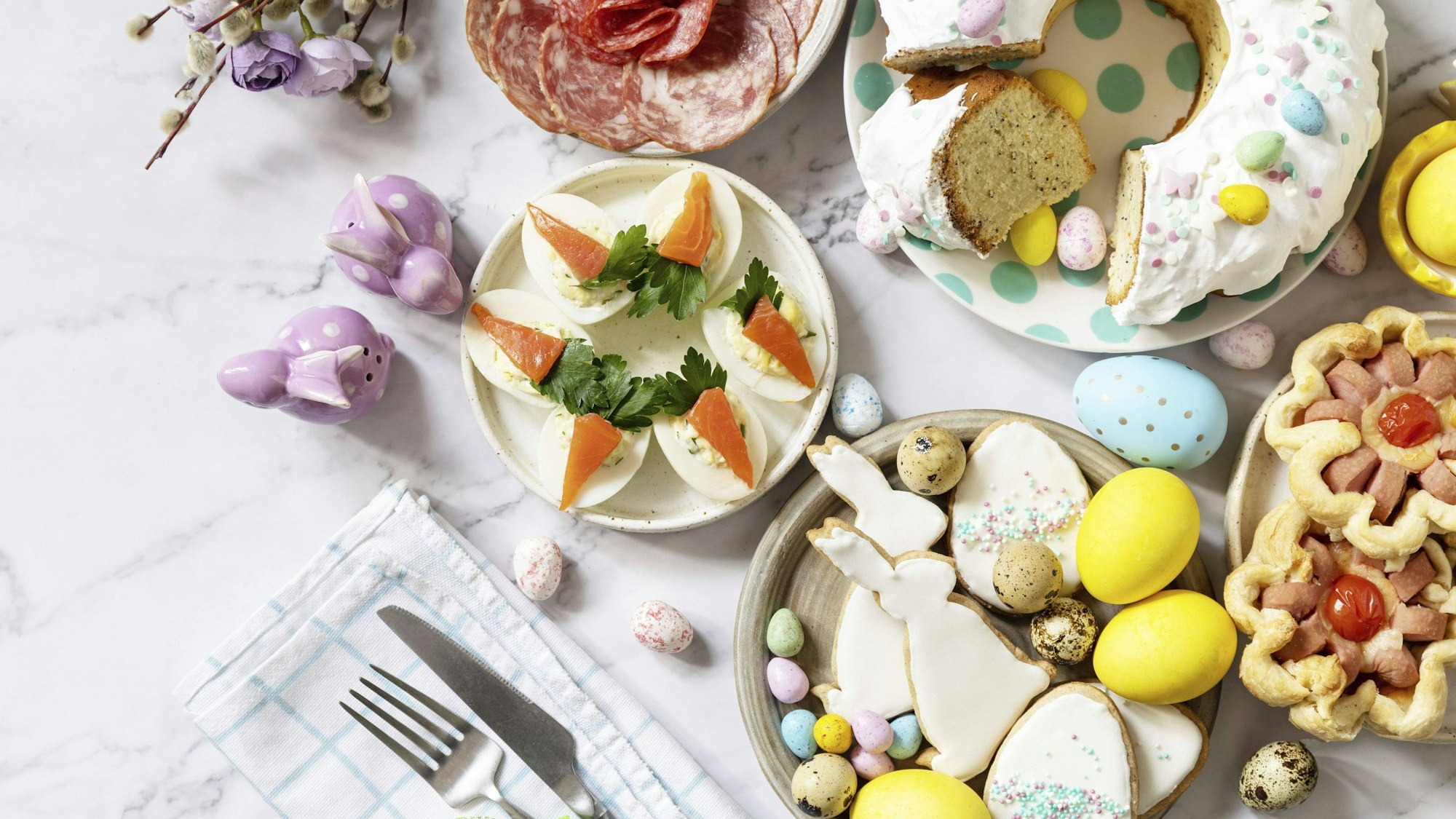 Ostertischszene mit einer Auswahl an Backwaren, Desserts, gefüllten Eiern und gefärbten Eiern auf einem Marmorhintergrund. Ansicht von oben.