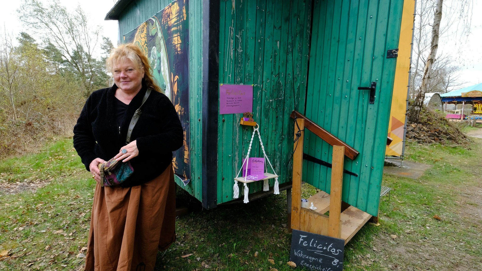Eine Frau mit roten Haaren und einem langen braunen Rock steht vor einem aus grünen Holzbrettern gezimmerten Schäferwagen.