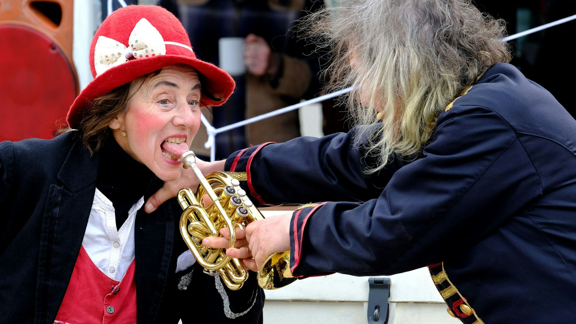 Ein Artistenpaar beim Auftritt: Der Frau, die einen roten Hut trägt, klebt scheinbar eine Trompete an der Zunge fest, ihr Partner versucht das Instrument zu lösen.