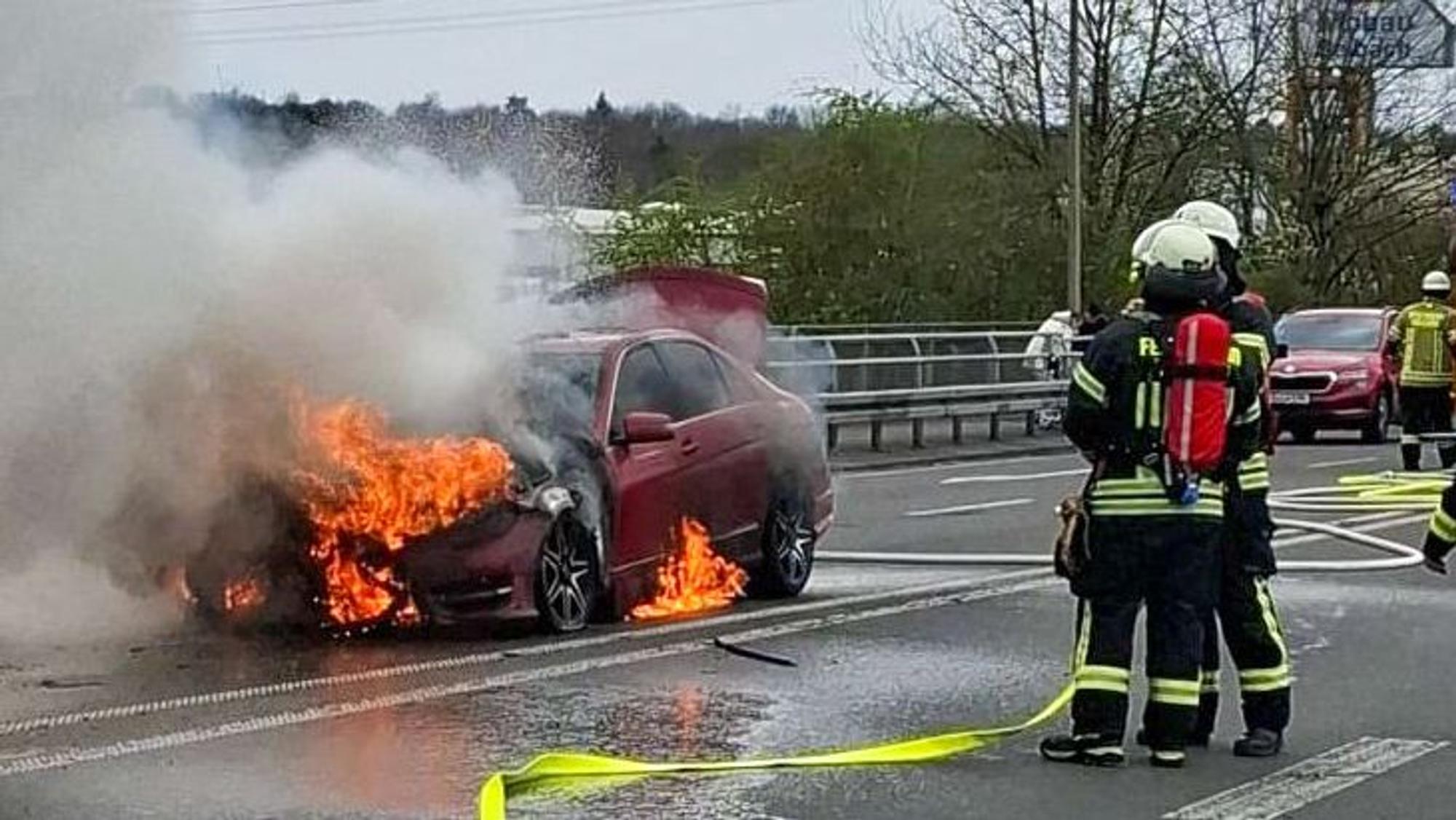 Feuerwehrleute stehen neben einem brennenden Auto.