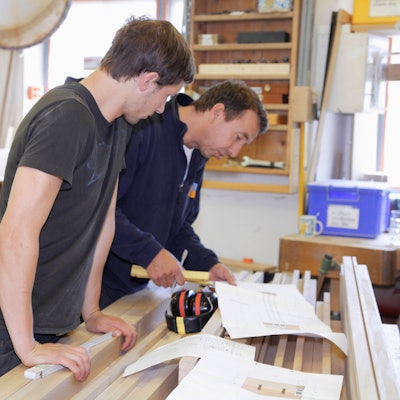 Zwei Männer messen Holz zusammen in einer Werkstatt aus.