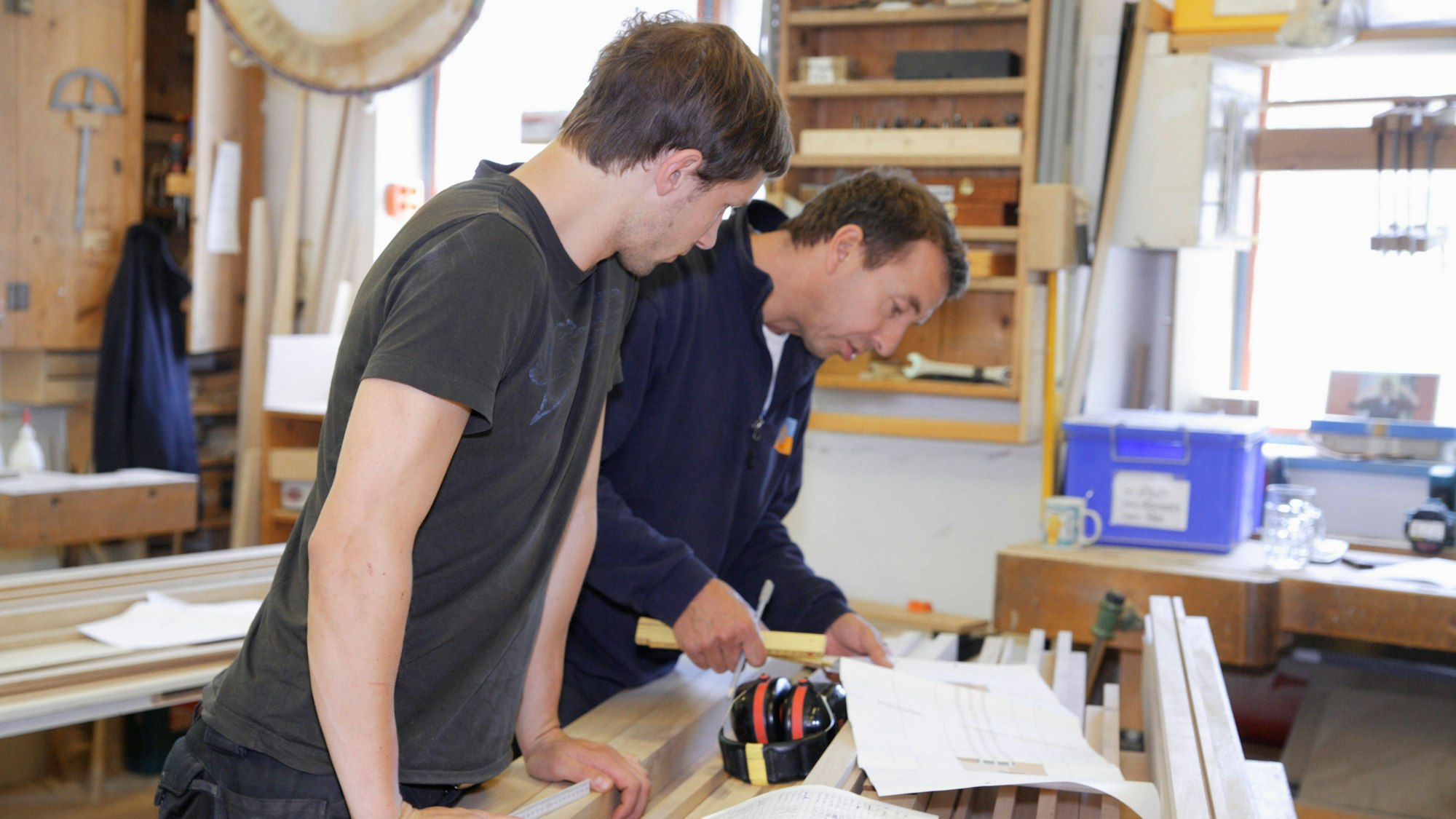 Zwei Männer messen Holz zusammen in einer Werkstatt aus.