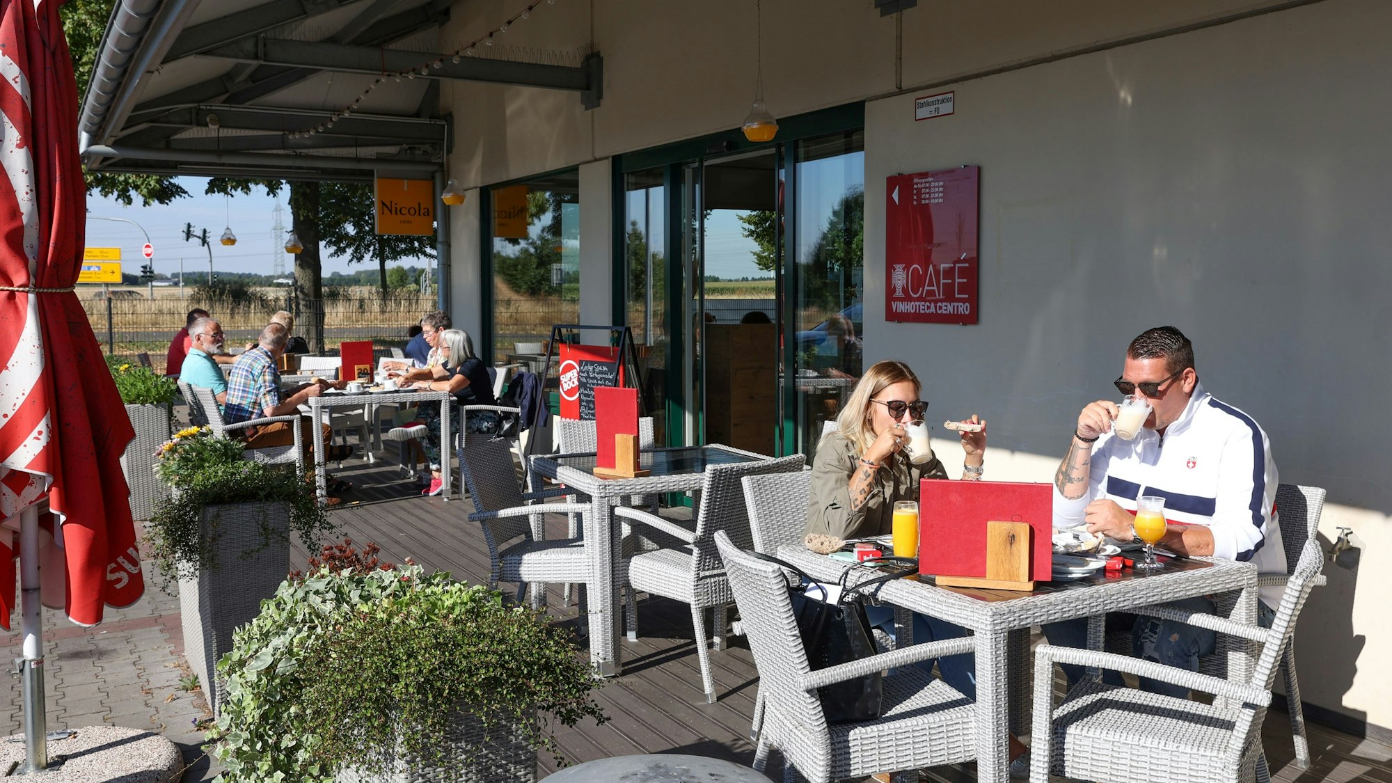 Das Vinhoteca Centro in Rommerskirchen von außen. Eine Terrasse des Cafés bietet Außengastronmie, Menschen sitzen an Tischen und essen und trinken.