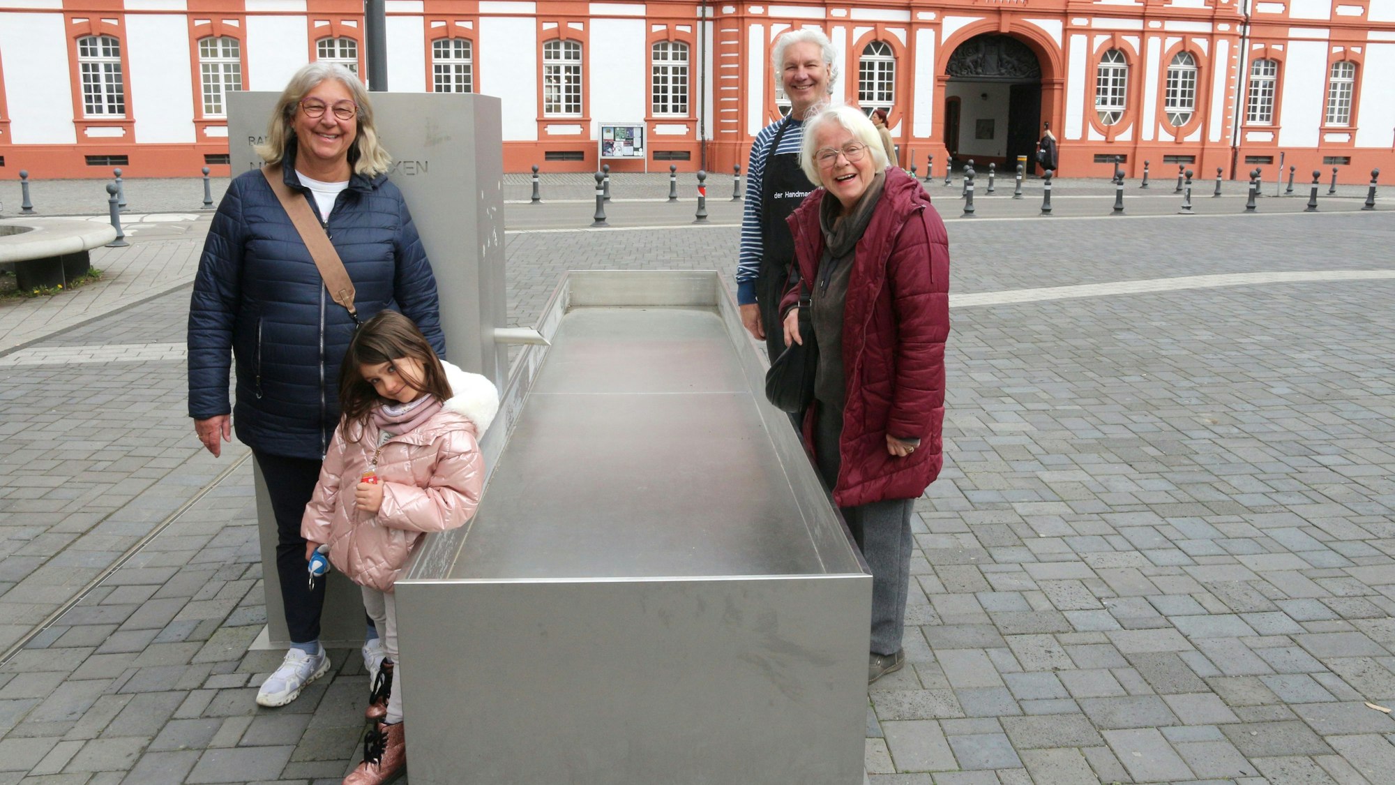 Drei Erwachsene und ein Kind stehen an einem Brunnen, Wasser sprudelt nicht.