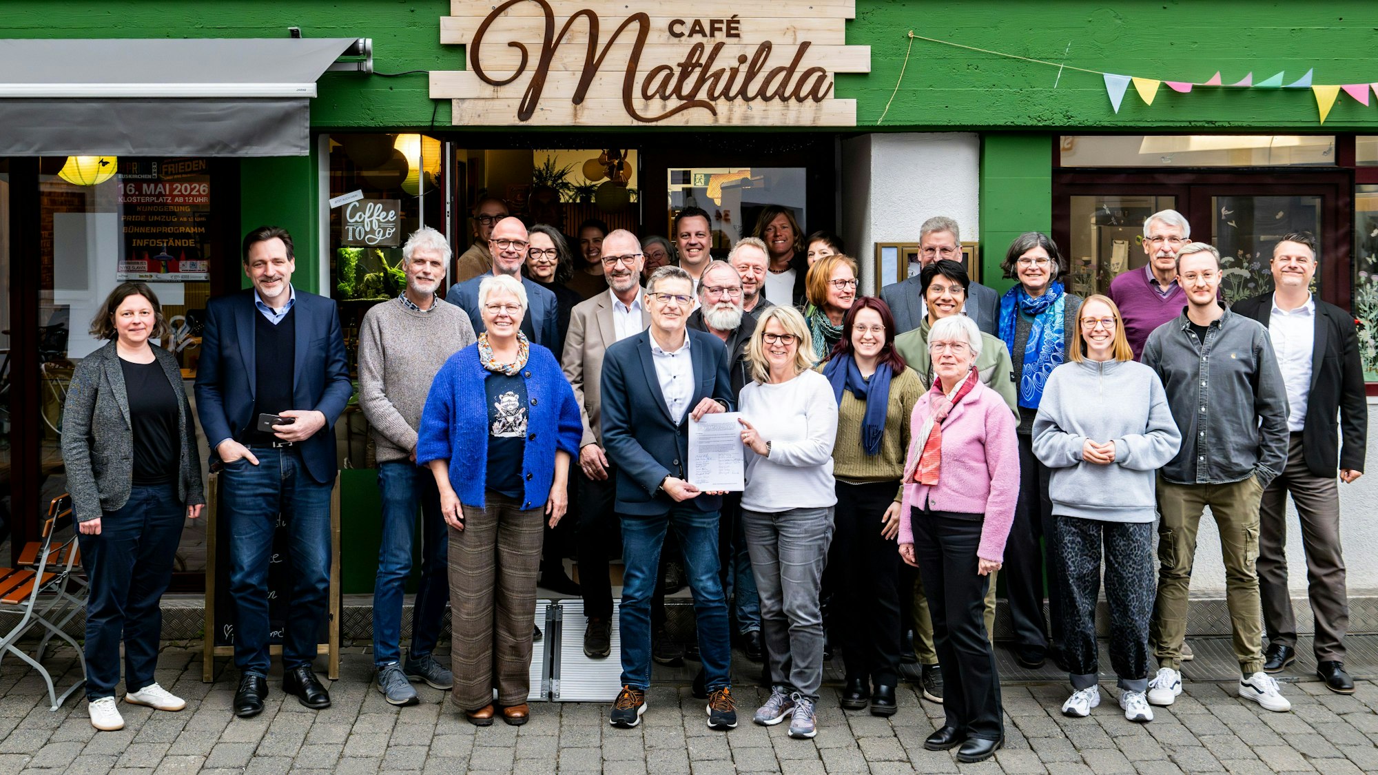 Eine Gruppe Menschen steht vor einem Haus mit grüner Fassade, an dem das Schild „Café Mathilda“ angebracht ist.