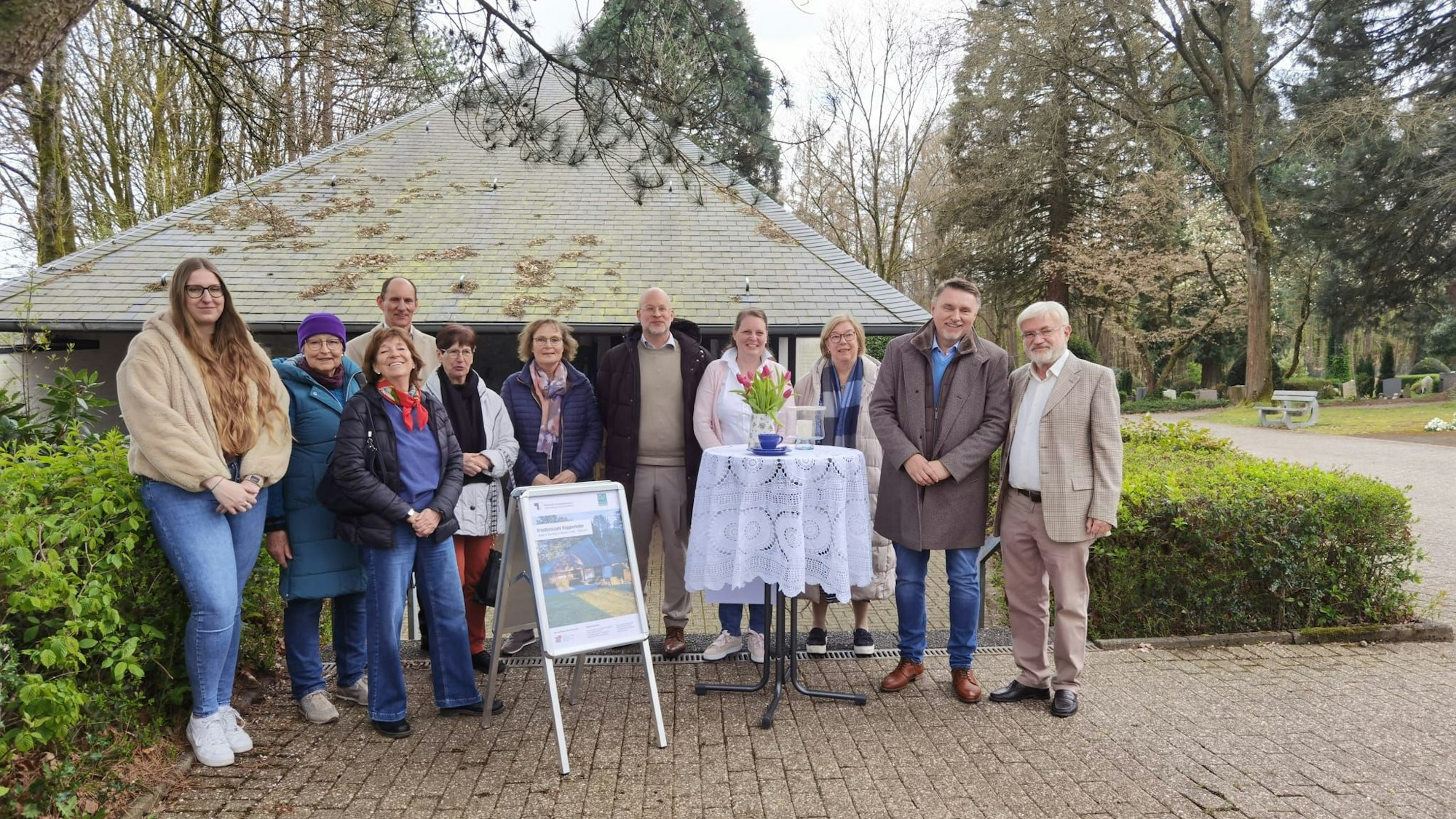 Eine Gruppe steht auf einem Friedhof vor einer Trauerhalle und lächelt.
