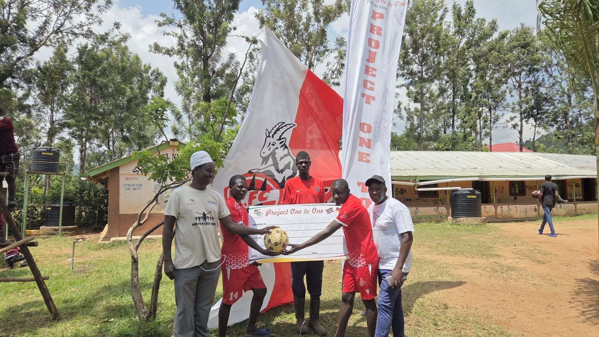 Der FC Cologne Kisumu wird eine Spende des Frechener Vereins „Project One to One“ überreicht, im Hintergrund die Flagge des 1. FC Köln.