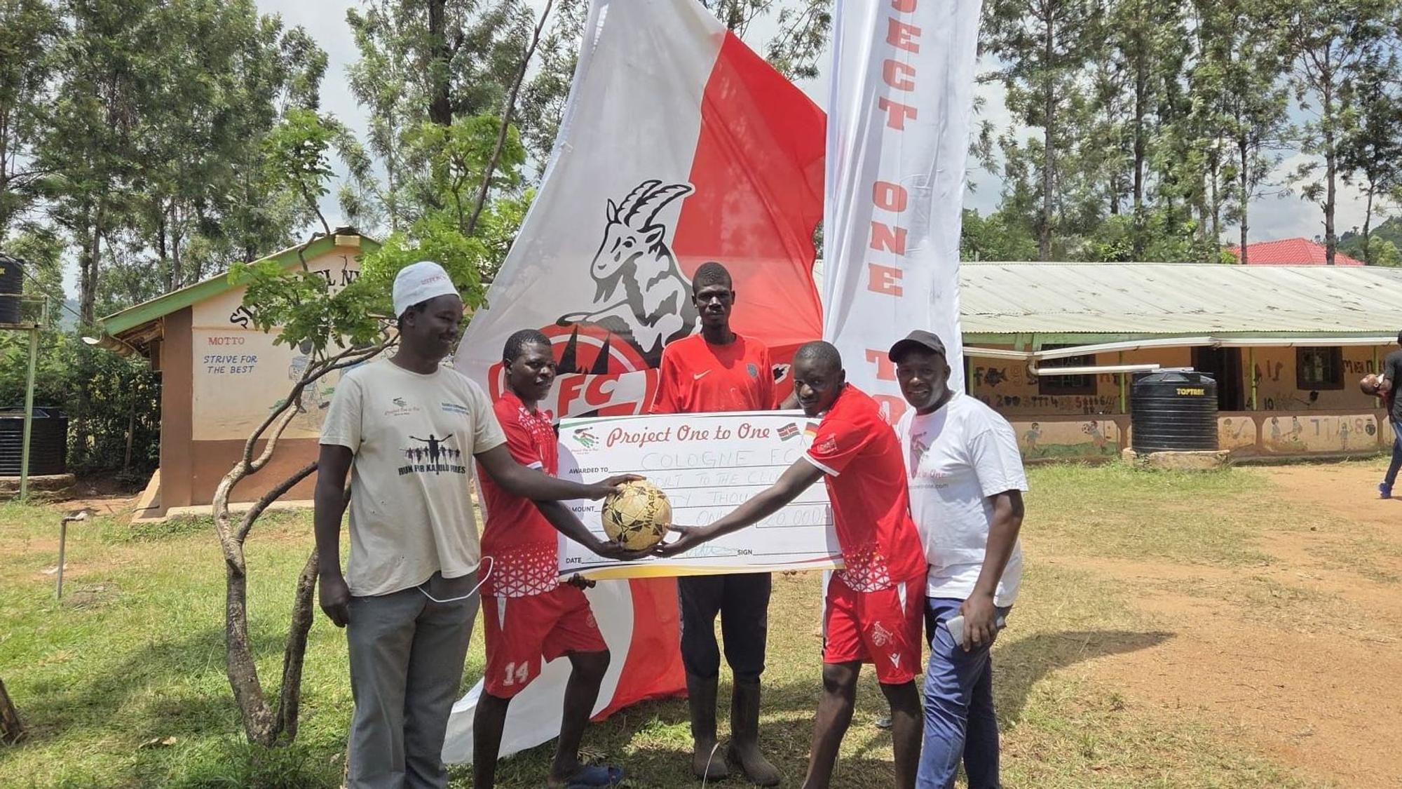 Der FC Cologne Kisumu wird eine Spende des Frechener Vereins „Project One to One“ überreicht, im Hintergrund die Flagge des 1. FC Köln.