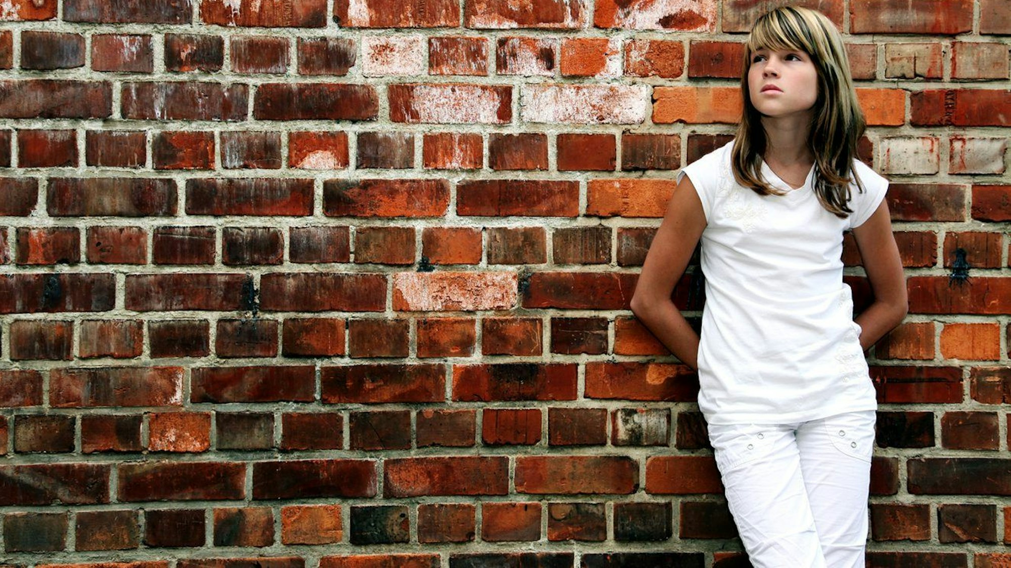 Ein weiß gekleidetes Mädchen steht vor einer Steinmauer und schaut traurig in die Luft.