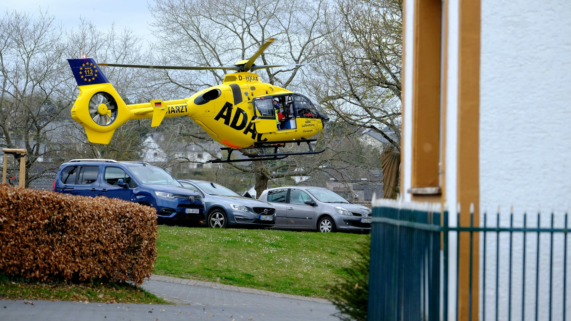Der gelbe ADAC-Rettungshubschrauber landet auf einer kleinen Grünfläche, dahinter stehen geparkte Autos.
