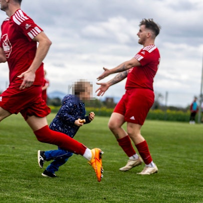 Ein Kind läuft, mit der Wurst in der Hand, übers Spielfeld und zwischen zwei Verteidigern hindurch. Einer von ihnen weist gestenreich auf den Flitzer hin.