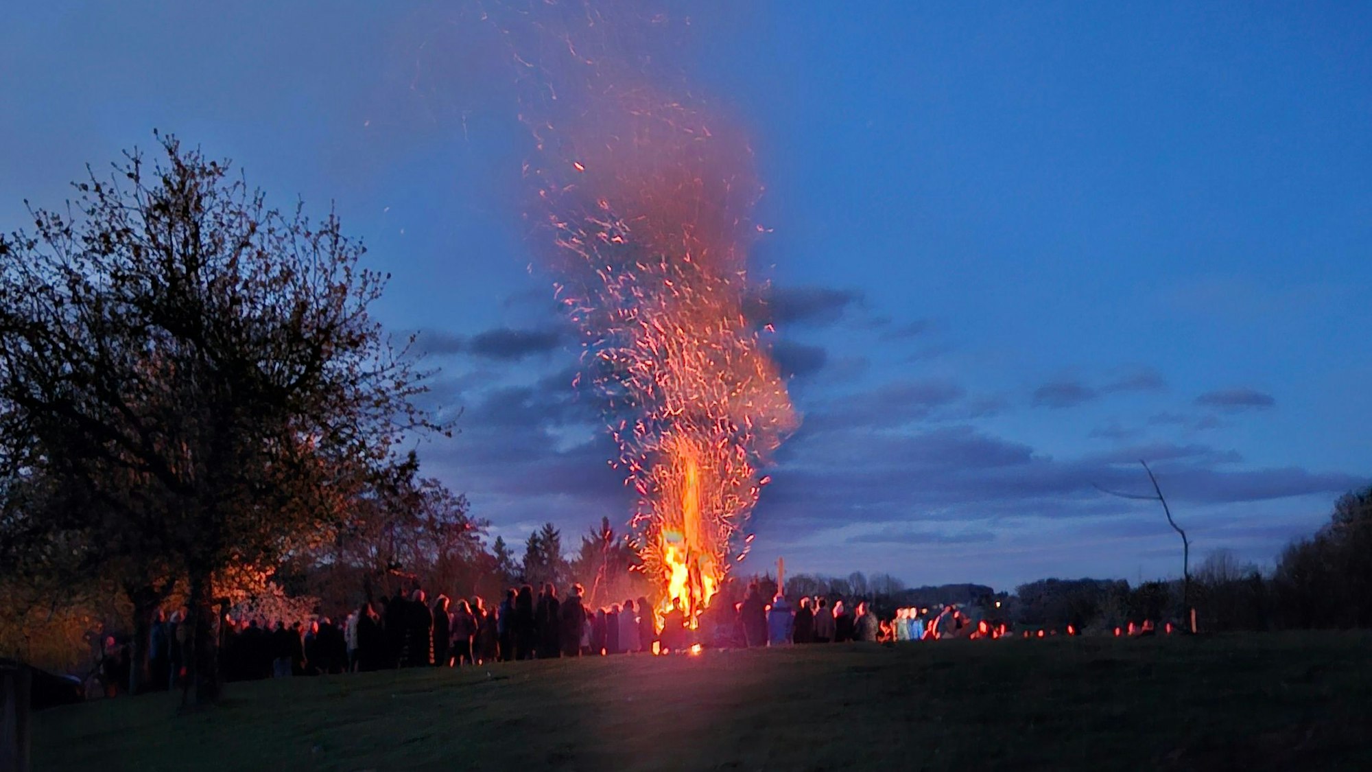 Aus der Ferne: Ein großes Feuer vor einem dunklen Nachhimmel, darum herum stehen Menschen