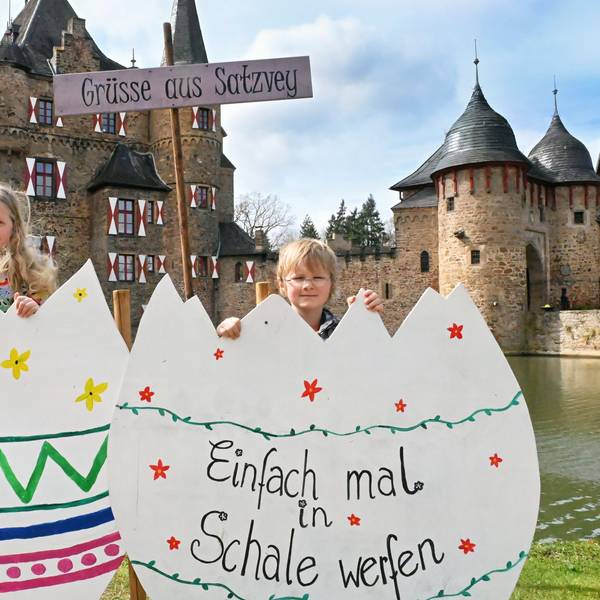 Die beiden Kinder stehen hinter zwei großen Pappeiern. Auf einem steht zu lesen: „Einfach mal in Schale werfen“. Hinter ihnen ein Schild mit der Aufschrift „Grüße aus Satzvey“, dahinter die Burg.