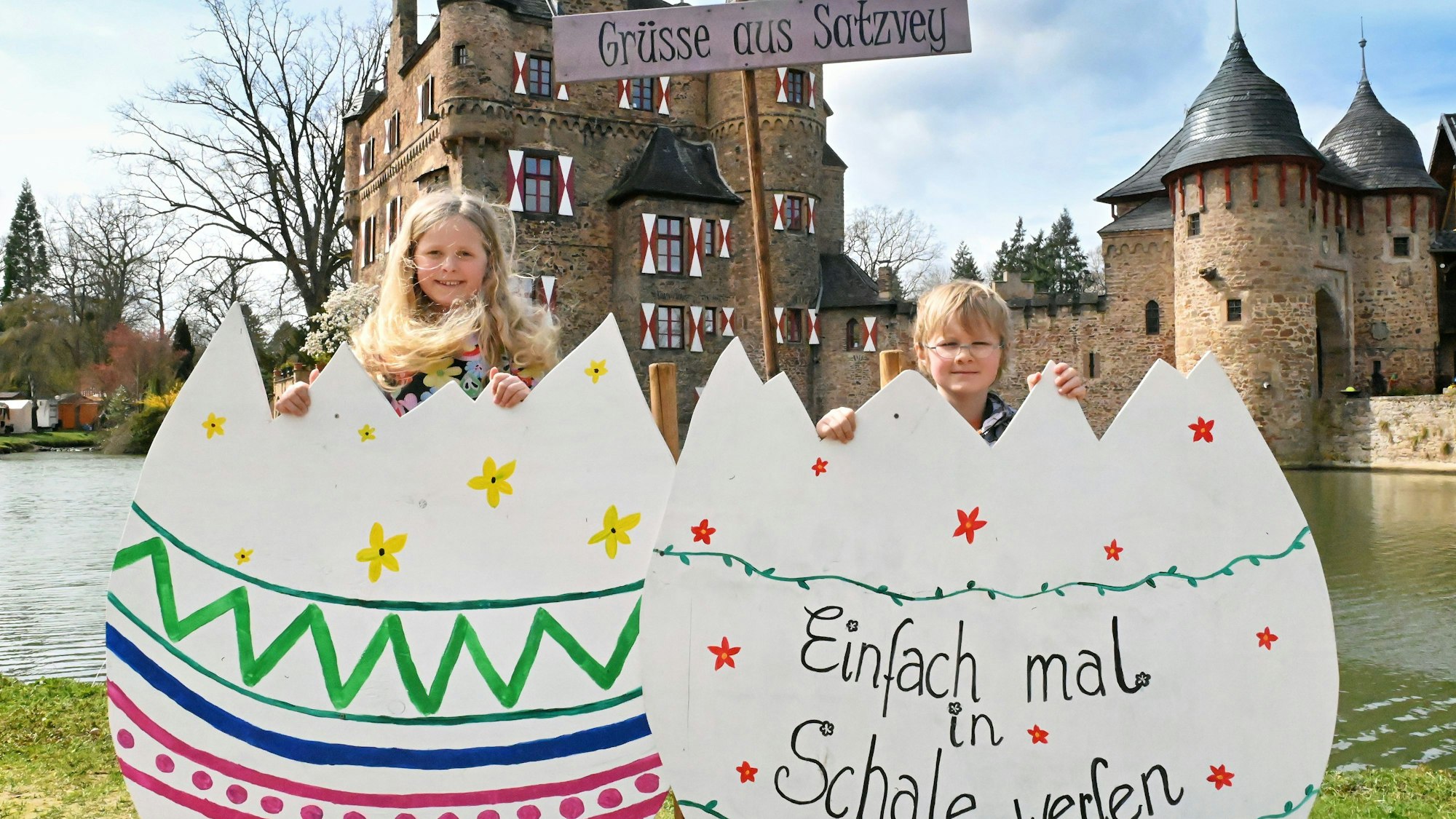 Die beiden Kinder stehen hinter zwei großen Pappeiern. Auf einem steht zu lesen: „Einfach mal in Schale werfen“. Hinter ihnen ein Schild mit der Aufschrift „Grüße aus Satzvey“, dahinter die Burg.