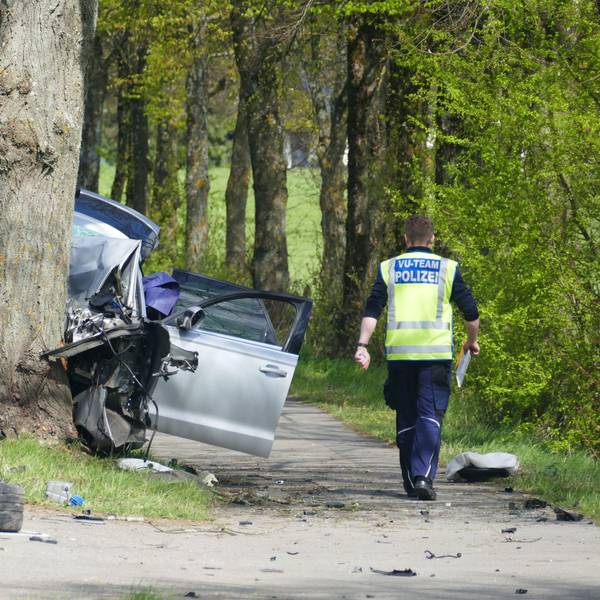 Rechts ein Polizist von hinten, in der Mitte ein Baumstamm, gegen den ein Pkw gefahren ist, herumliegende Trümmerteile