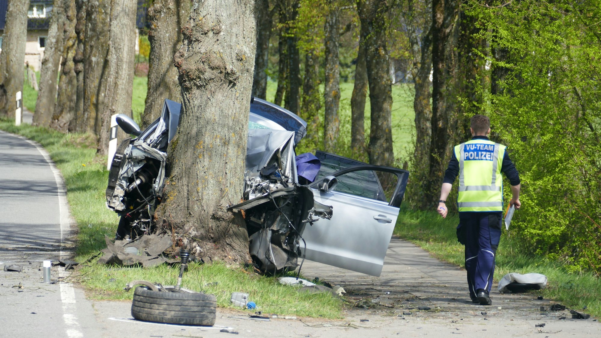 Rechts ein Polizist von hinten, in der Mitte ein Baumstamm, gegen den ein Pkw gefahren ist, herumliegende Trümmerteile