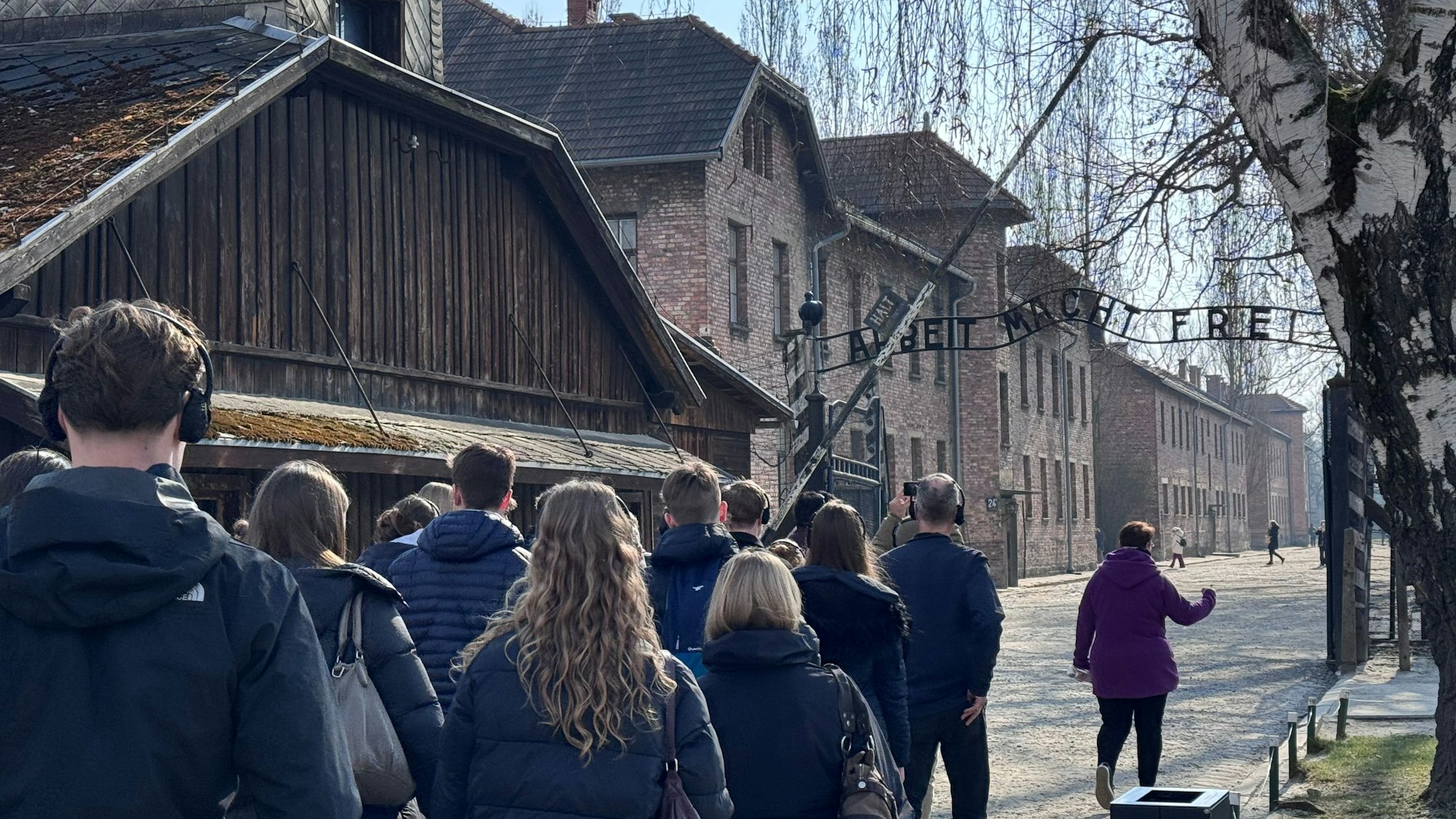 Eine Gruppe läuft durch ein Stammlager in Auschwitz unter einem Tor mit der Aufschrift "Arbeit macht frei" durch.