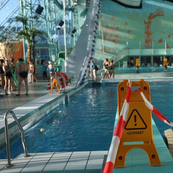 Ein Schild mit der Aufschrift „geperrt“ steht auf einem Sprungbrett. Im Hintergrund sieht man ein Schwimmbecken, um das Menschen laufen.